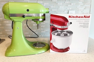 A kitchenaid mixer with the ice cream attachment still in the box next to it.