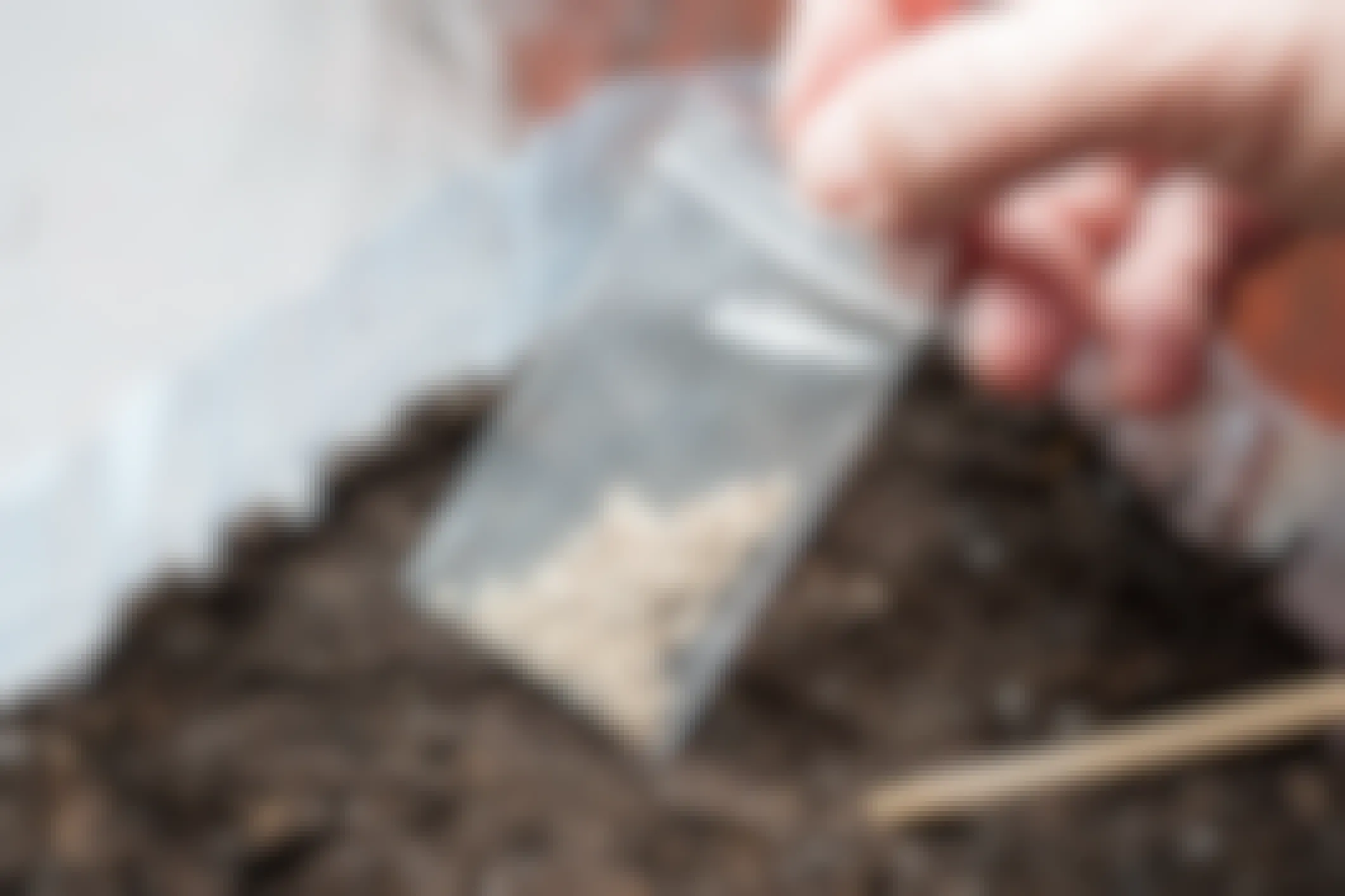 A person's hand holding a small bag of seeds above a container of soil.