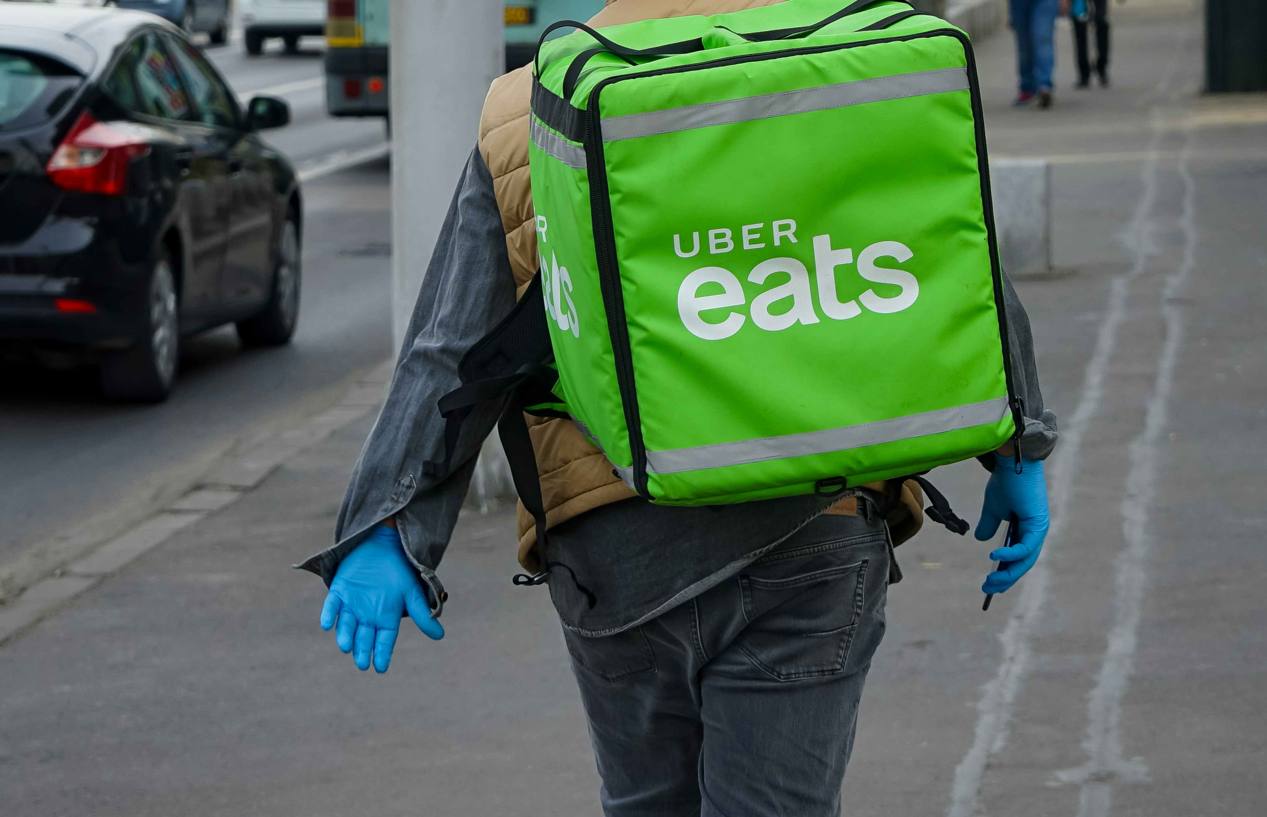 An Uber Eats worker walking down the sidewalk wearing an Uber Eats meal delivery backpack.