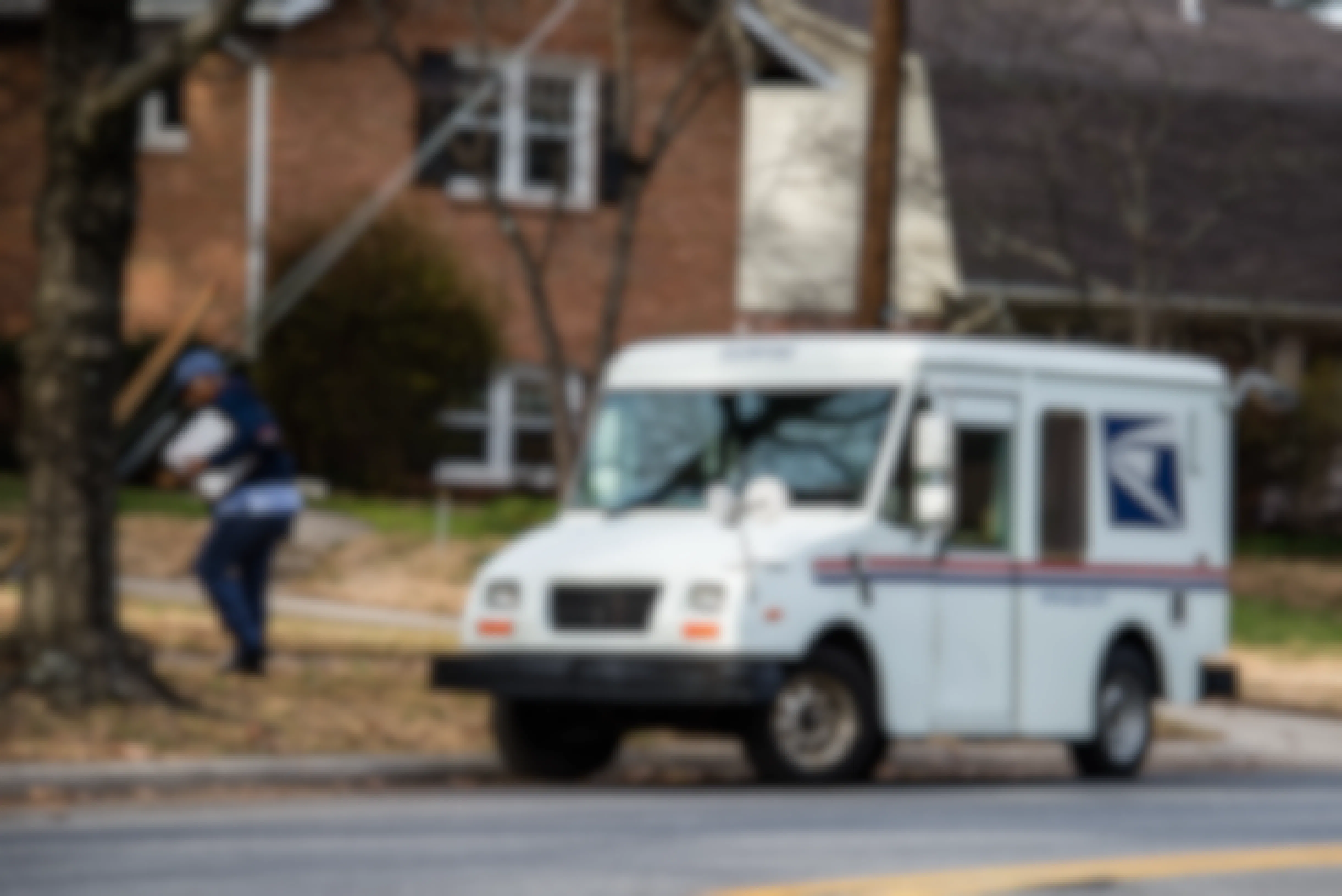 A USPS mail truck pulled up to a curb and a postal worker walking toward a house, carrying a package.