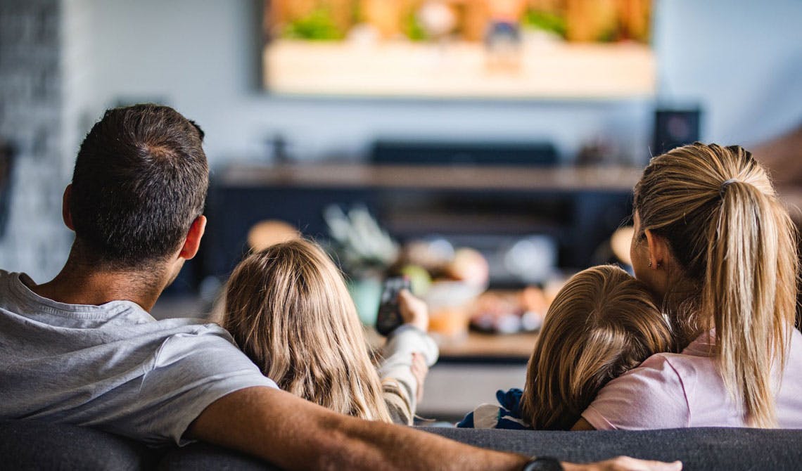 A family sitting on a couch facing away toward a TV mounted on the wall; one of the children is raising the remote up, pointing at the TV.