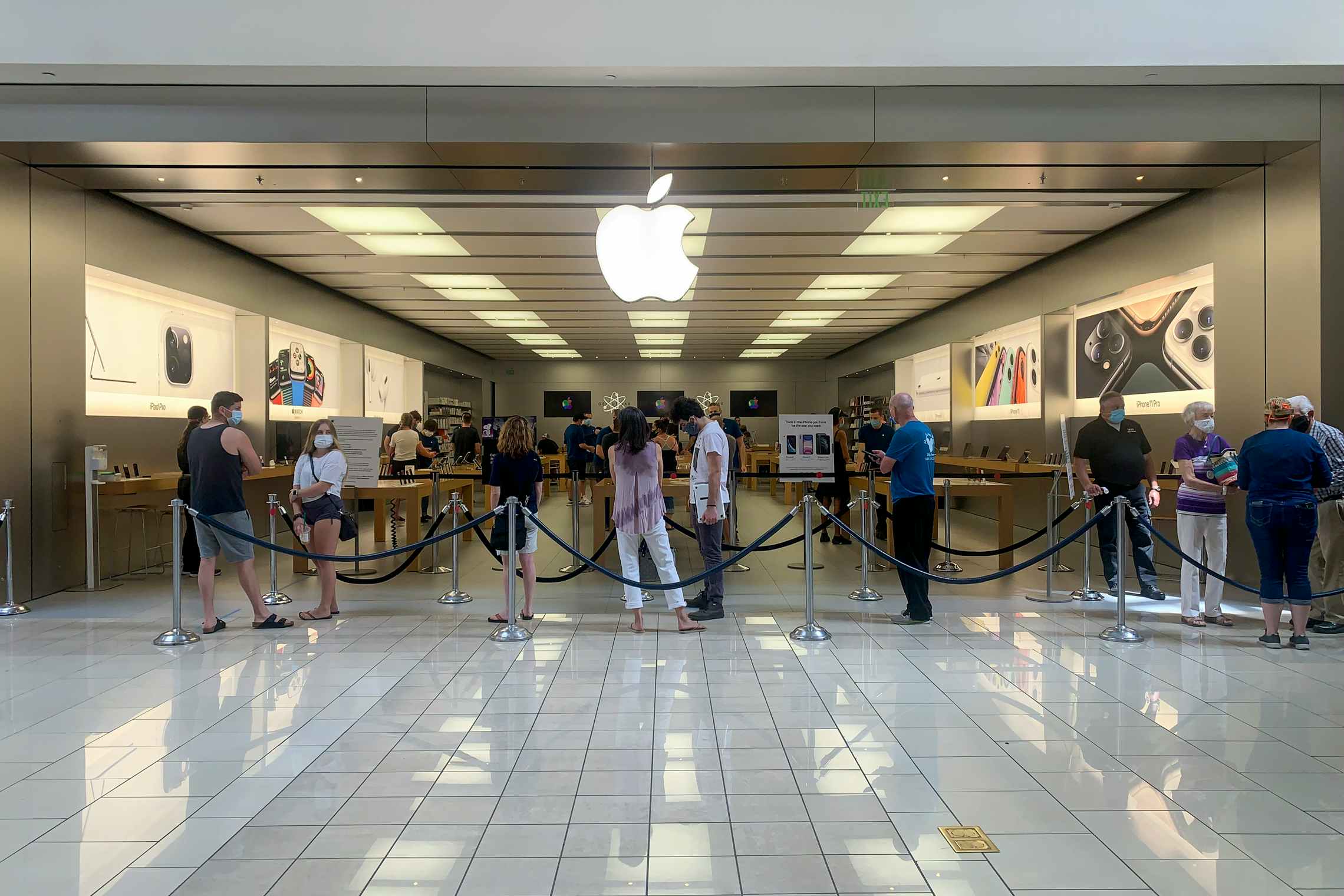 Customers wearing masks waiting in like to enter the Apple store inside a mall.