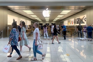 Customers wearing masks waiting in line to enter the Apple store inside a mall.