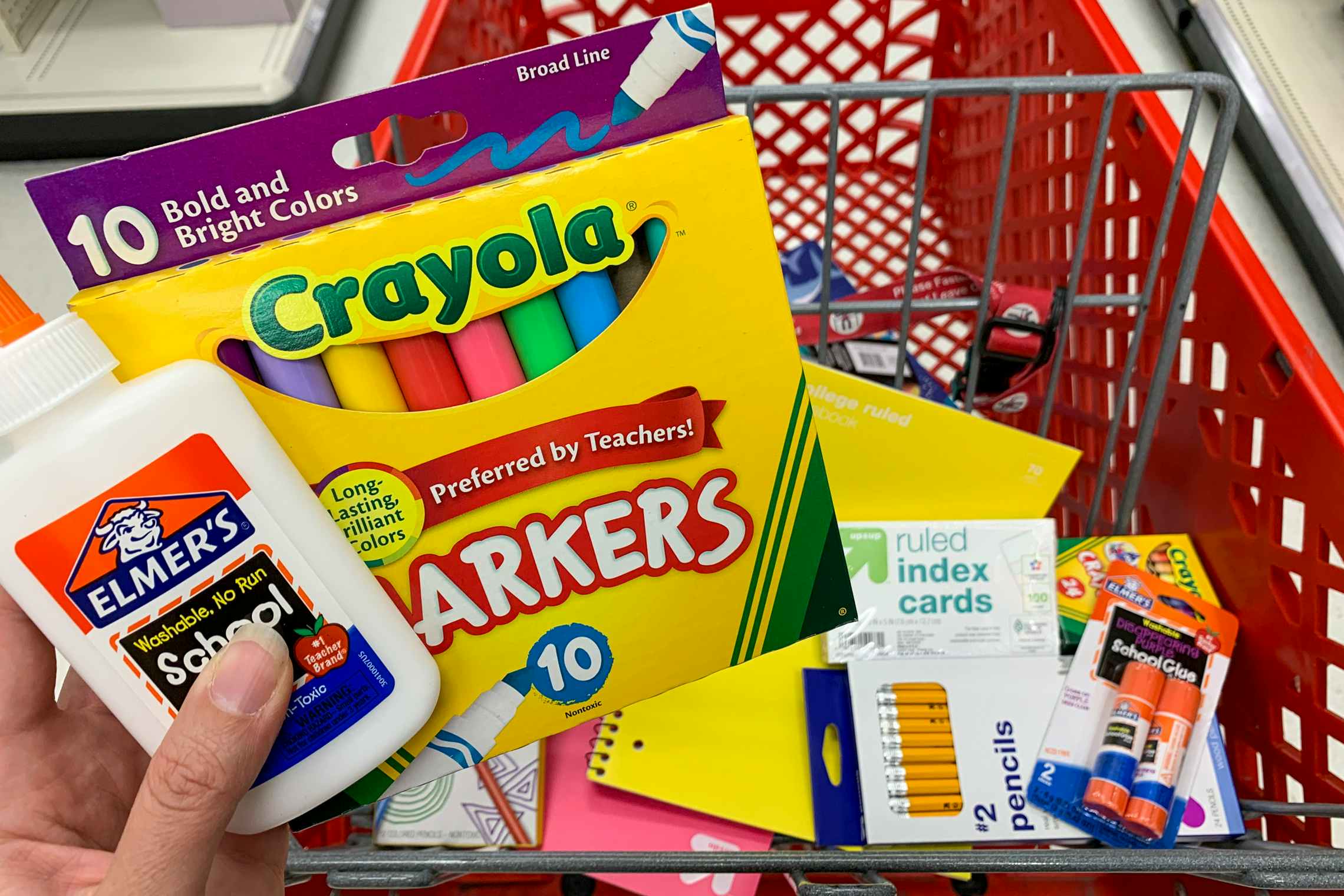 A person's hand holding Crayola markers and Elmer's glue above a Target shopping cart basket filled with other school supplies.