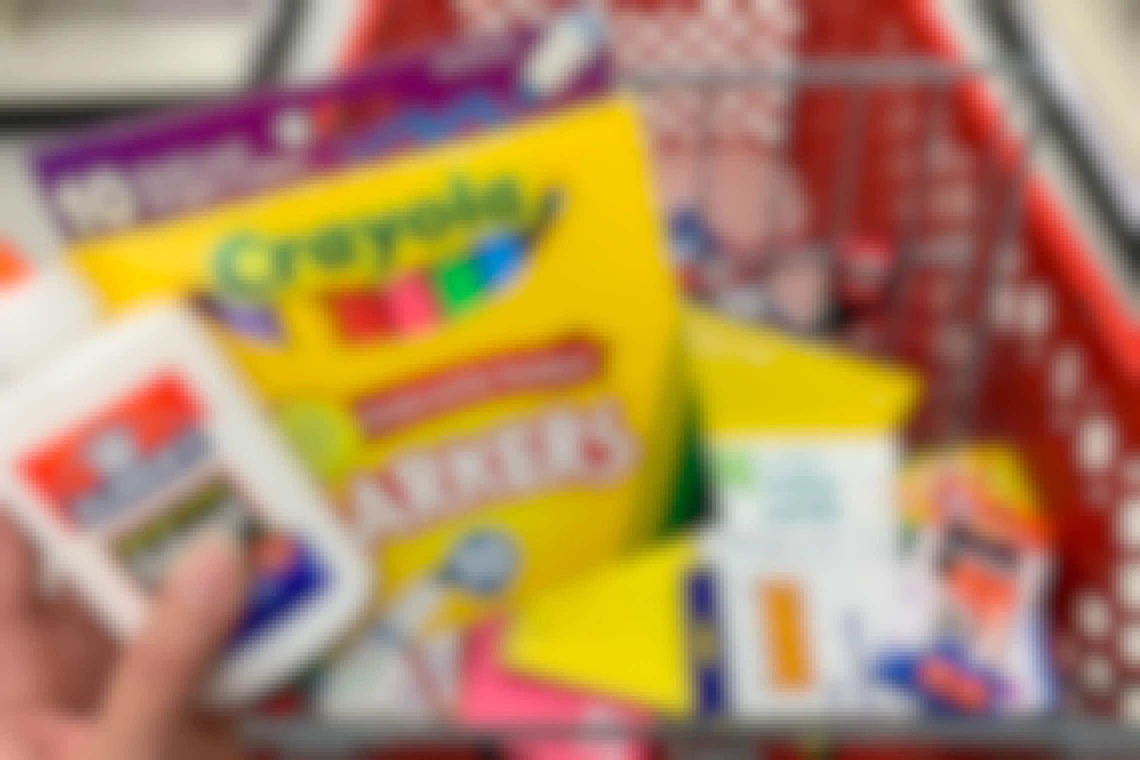 A person's hand holding Crayola markers and Elmer's glue above a Target shopping cart basket filled with other school supplies.