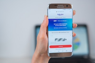 A woman holding a Samsung phone in the air with the Rakuten Ebates app pulled up on her screen.