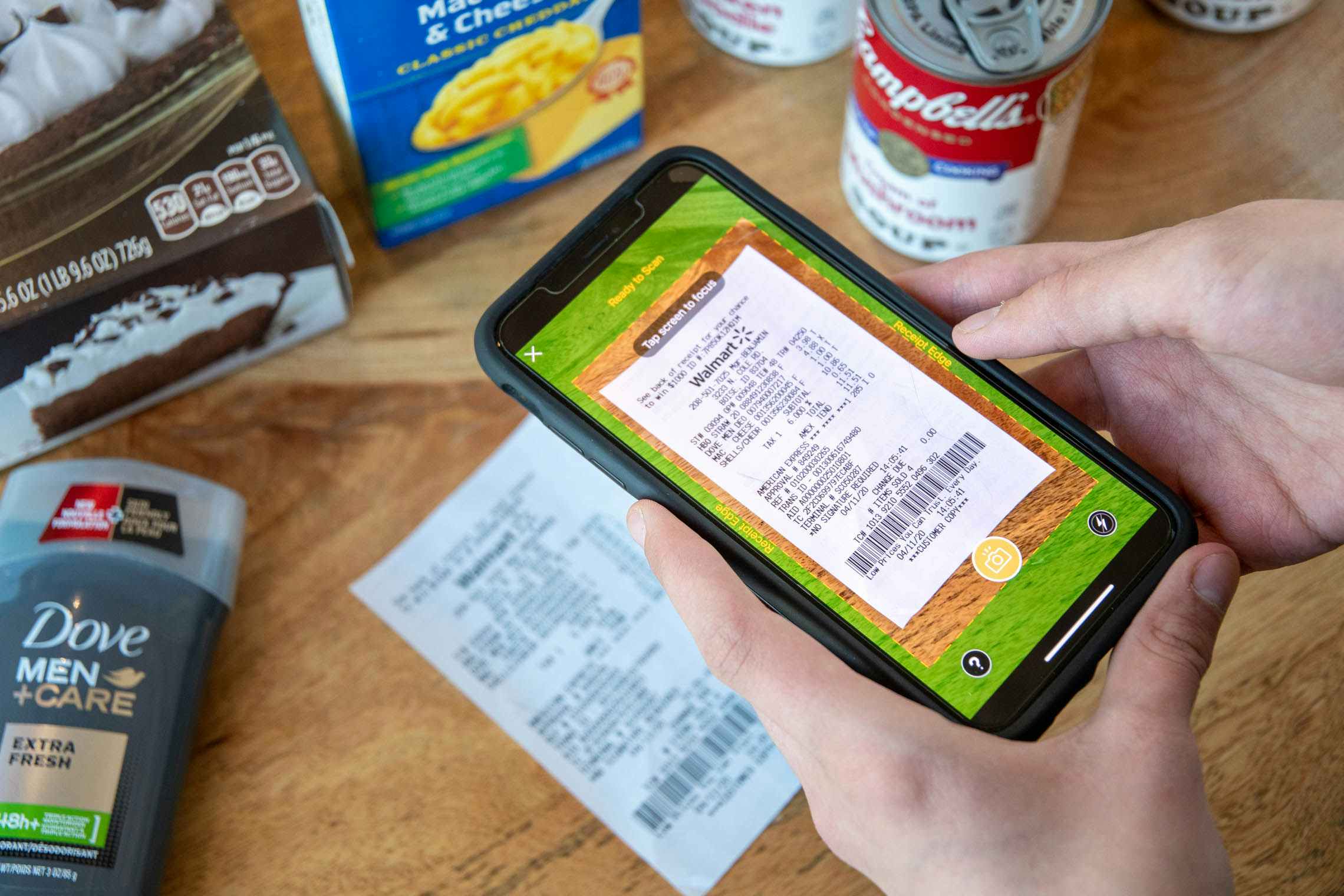 A person's hands holding an iPhone with the Fetch mobile app open to the receipt-scanning feature. The user is centering the phone's camera on a receipt that is laying on a table next to some grocery items.