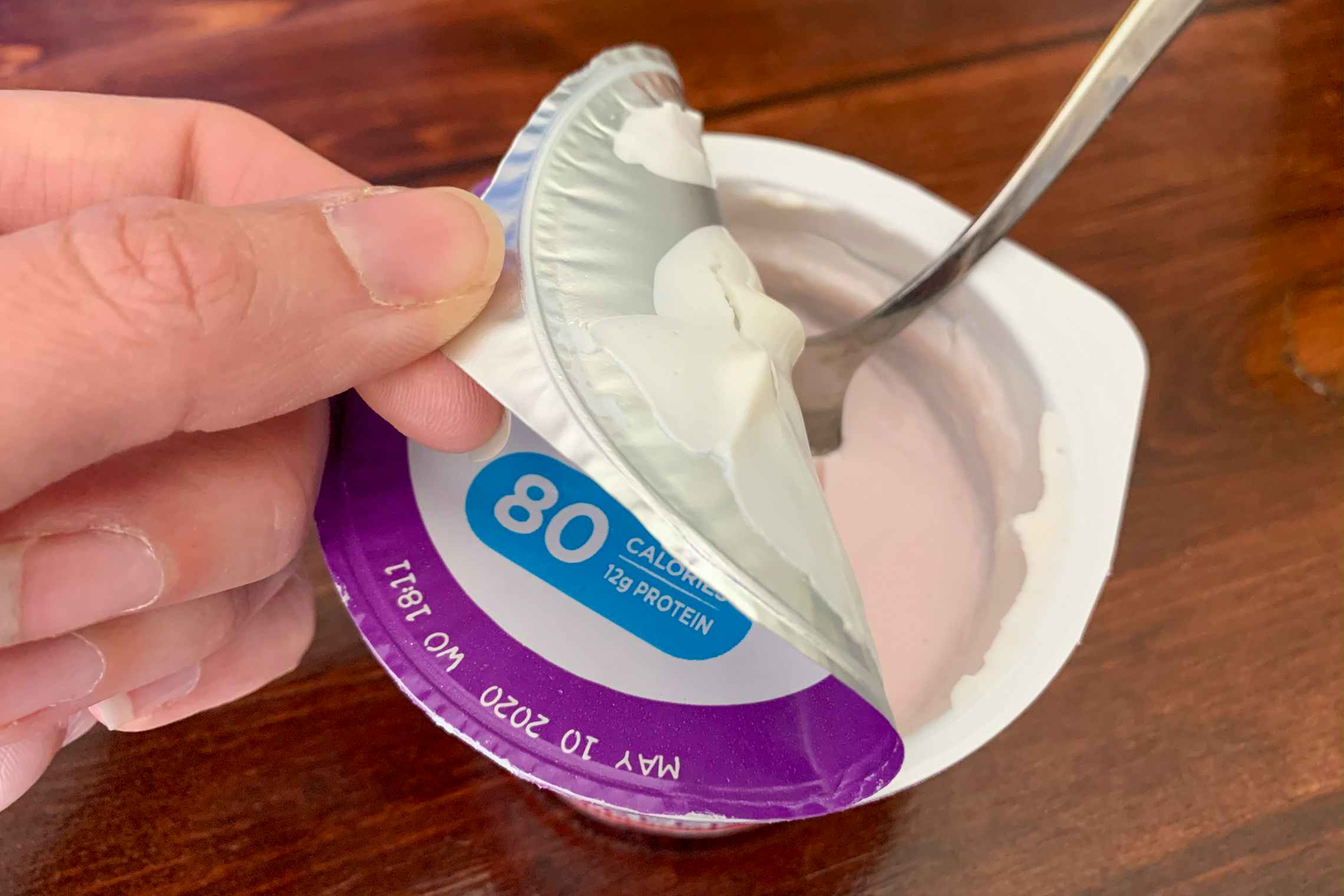 A spoon in a yogurt cup someone is pulling the metal lid off.