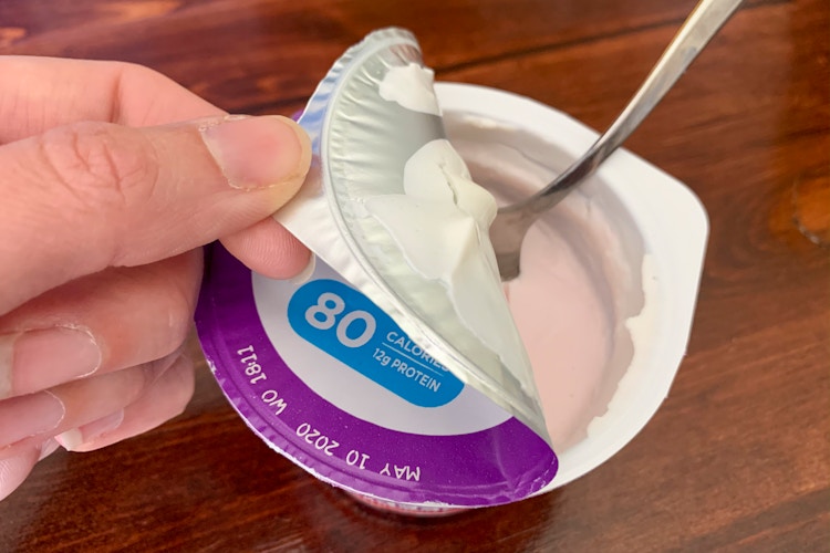 A spoon in a yogurt cup someone is pulling the metal lid off.