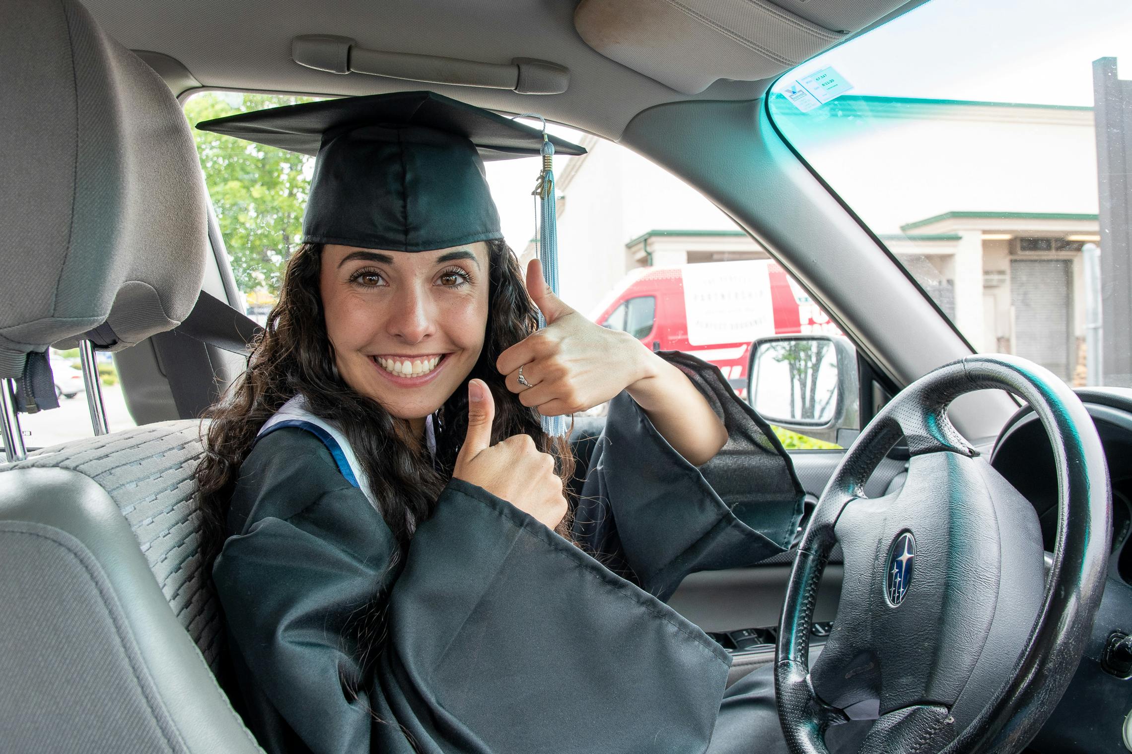 Woman in drivers seat of car, wearing a gradation cap and gown, holding two thumbs up.