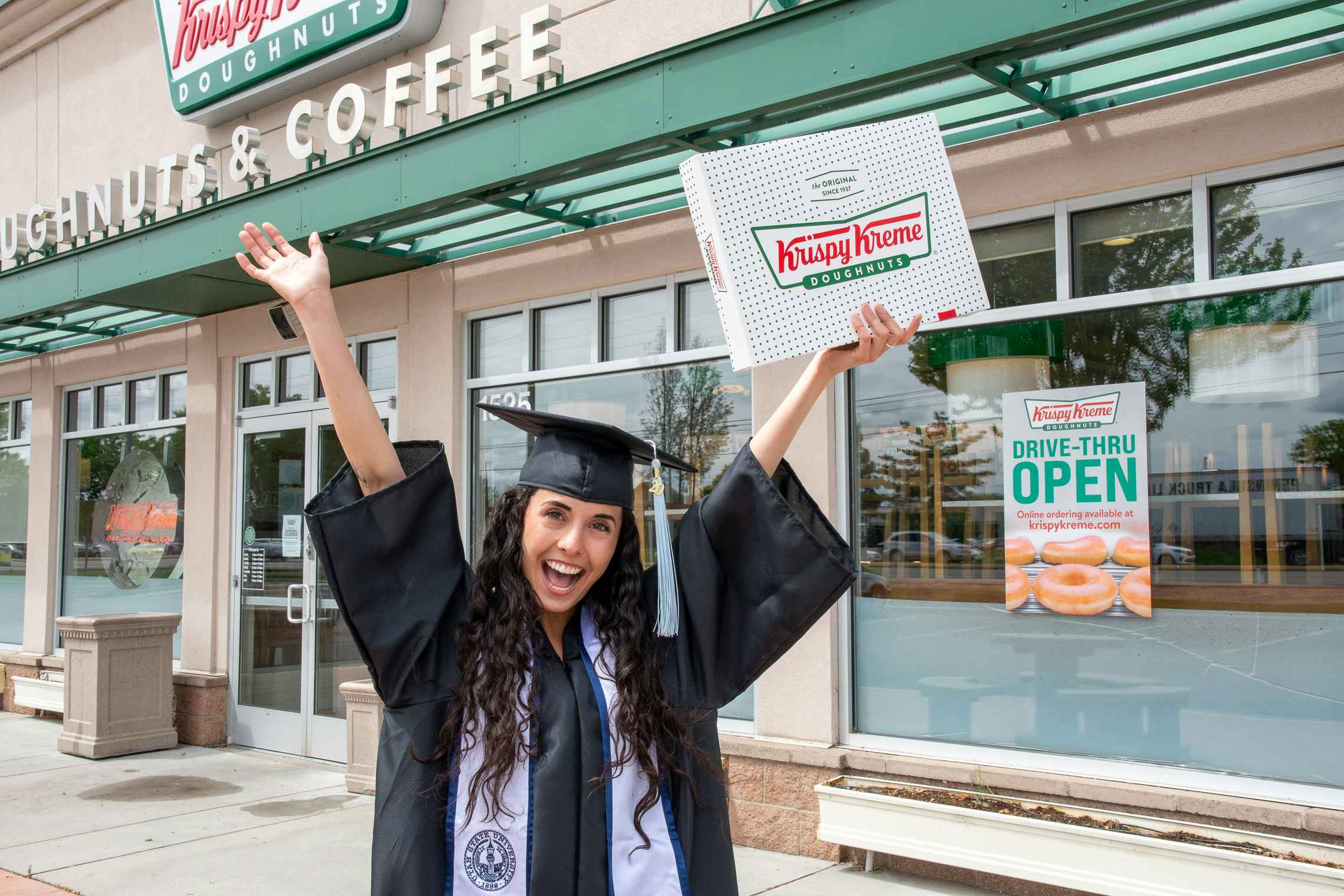 Woman in a graduation cap and gown holding up a box of a dozen doughnuts in front of a Krispy Kreme.