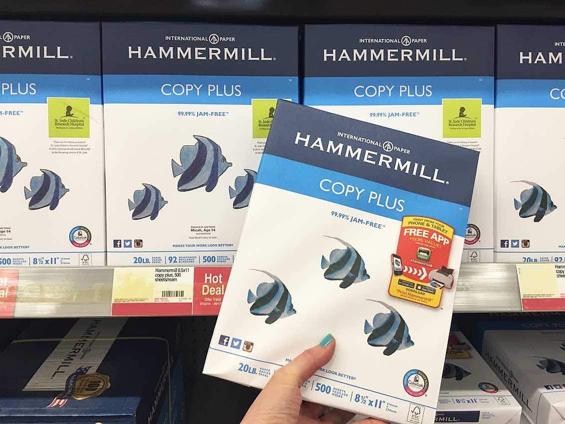 A person's hand holding a ream of Hammermill 8.5 x 11" paper in front of a shelf with more of the same product inside Staples.