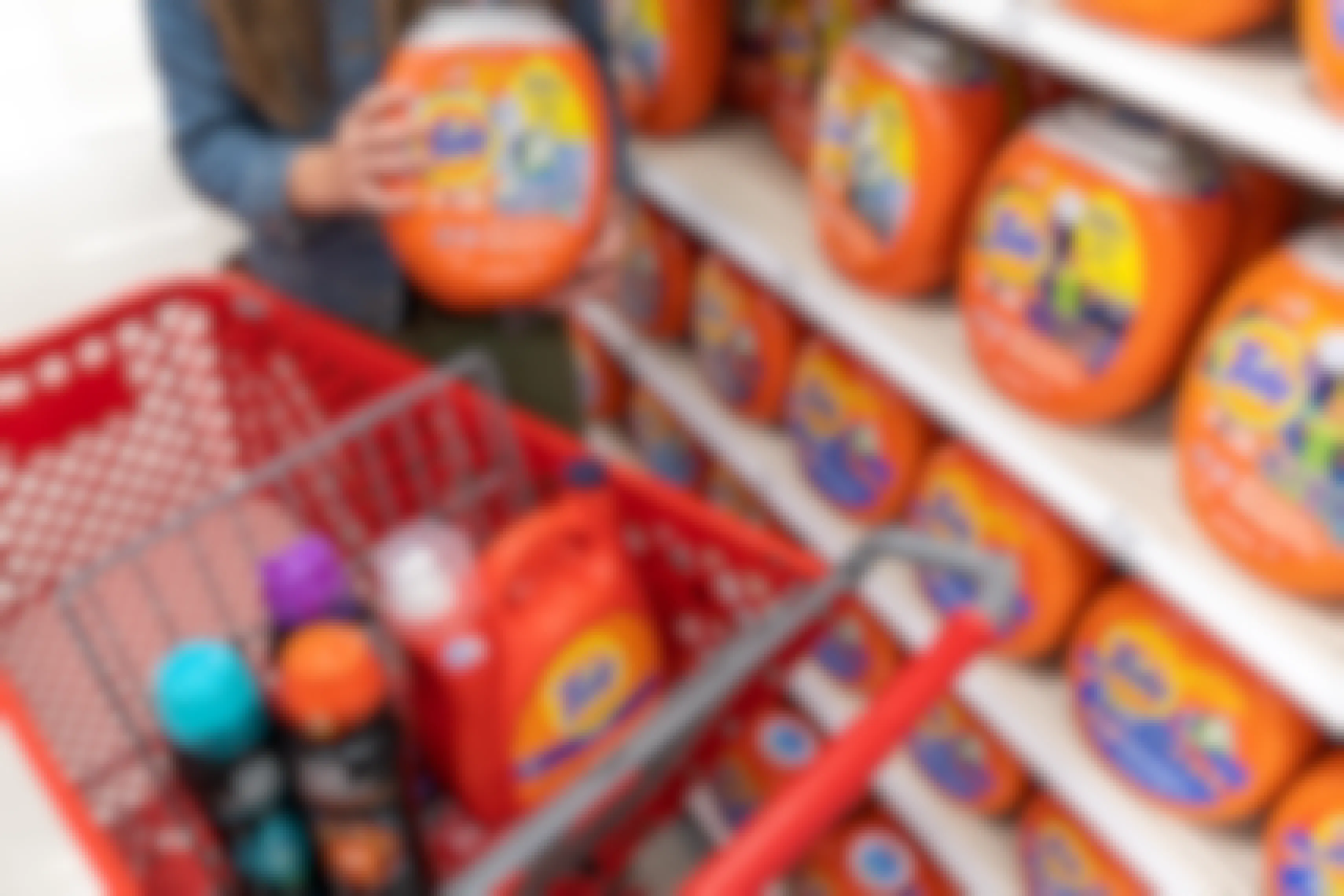 A woman standing behind a red Target shopping cart, holding a container of tide pods, with other laundry products in the basket.