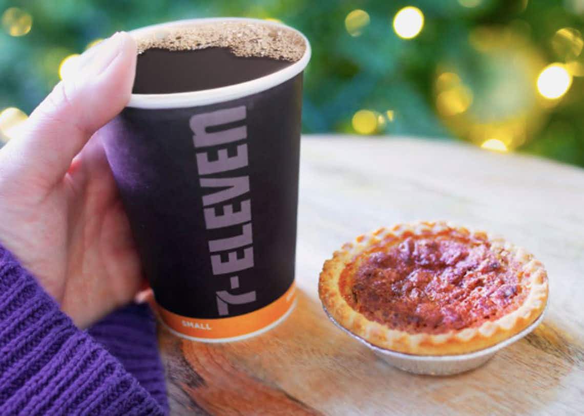 A person a holding 7-eleven cup filled with coffee next to a mini pumpkin pie.