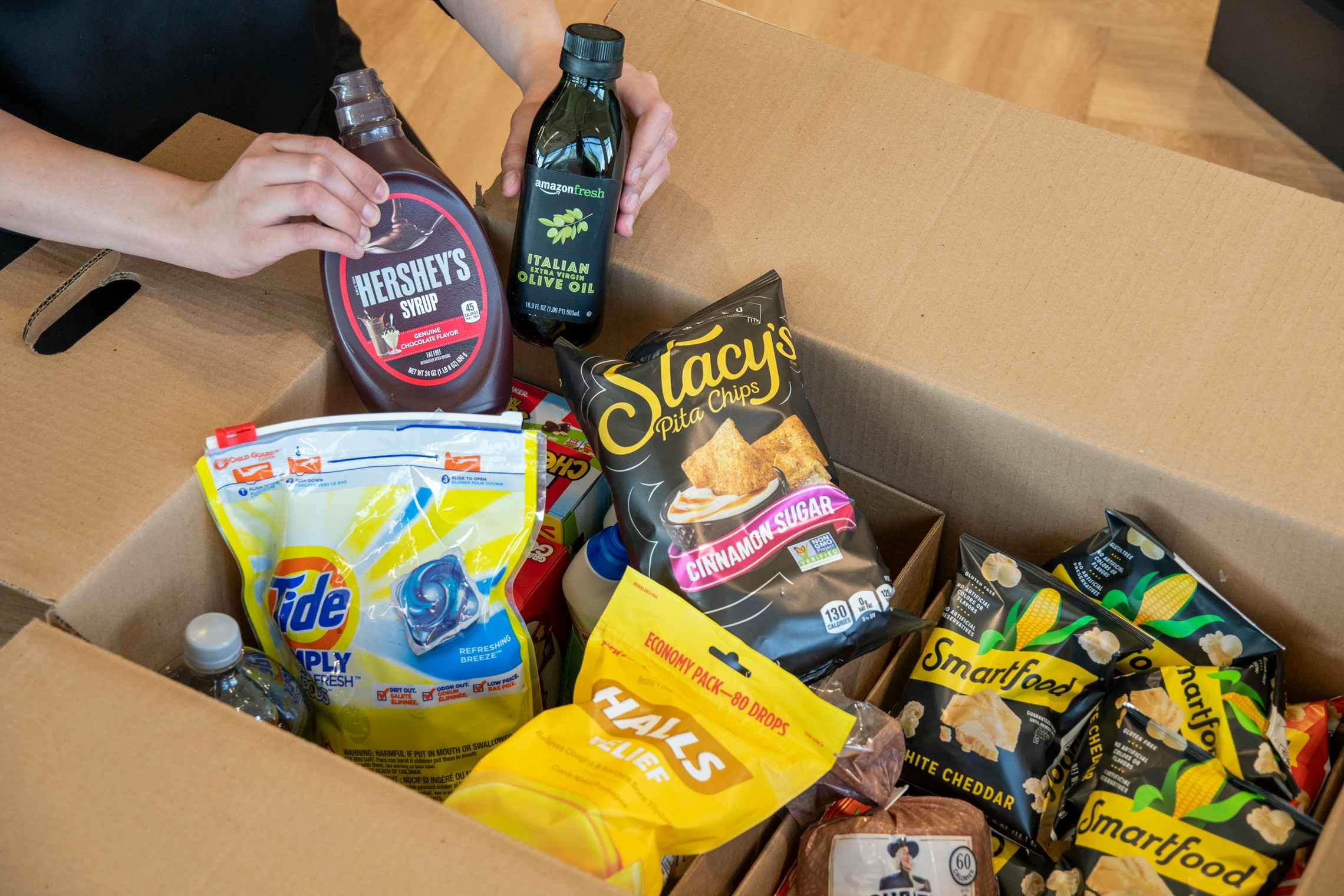 A person's hands holding up Hershey's chocolate syrup and Amazon Fresh Italian Olive Oil above a box of various groceries, including Stacy's Pita Chips, Halls cough drops, Smartfood Popcorn, and Tide Pods, ordered using Amazon Pantry.