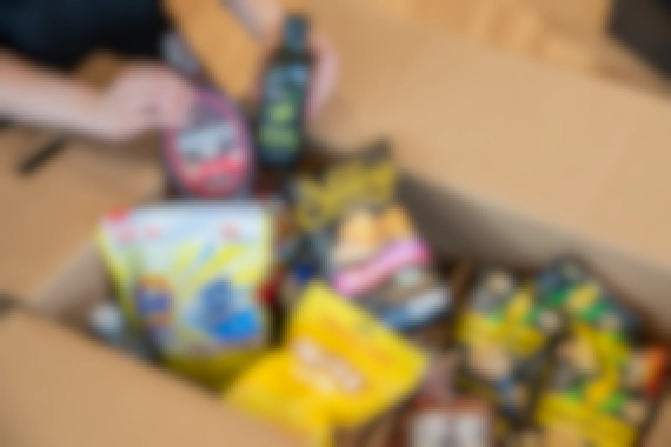 A person's hands holding up Hershey's chocolate syrup and Amazon Fresh Italian Olive Oil above a box of various groceries, including Stacy's Pita Chips, Halls cough drops, Smartfood Popcorn, and Tide Pods, ordered using Amazon Pantry.