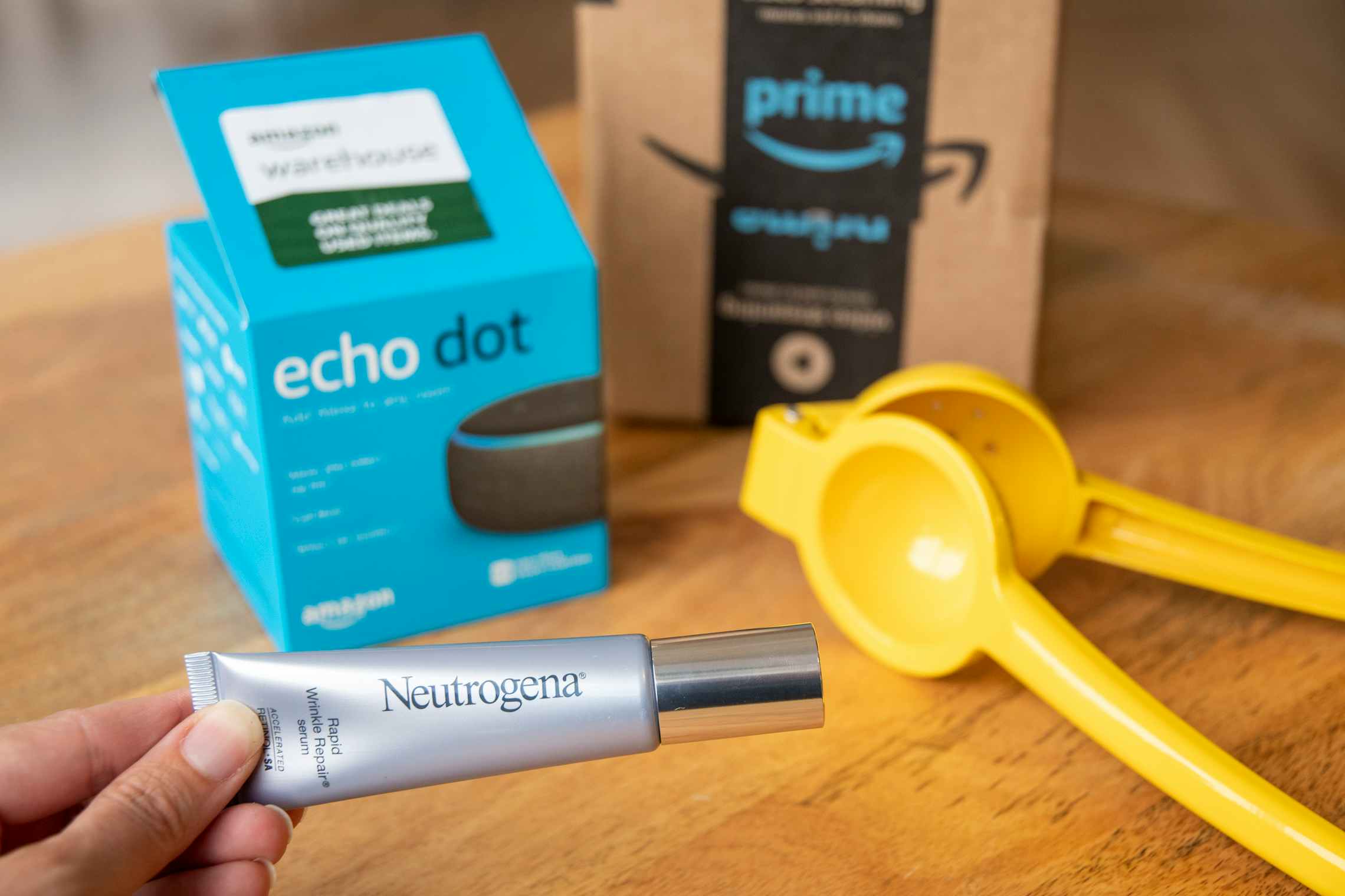 A person's hand holding a tube of Neutrogena Rapid Wrinkle Repair in front of an Echo Dot box, a kitchen utensil and an Amazon delivery box sitting on a table.