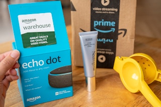 A person showing the Amazon Warehouse sticker on the lid of an Amazon Echo box on a table next to more products and a prime shipping box.