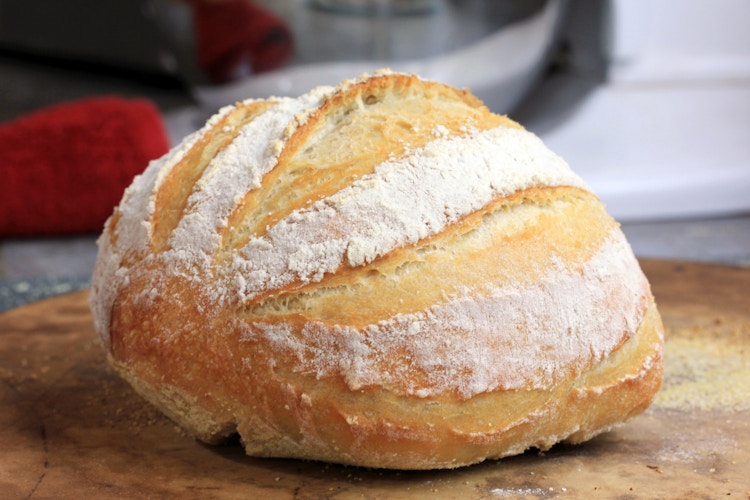 An round artisan loaf of bread with flour sprinkled on top sitting on a wooden surface.
