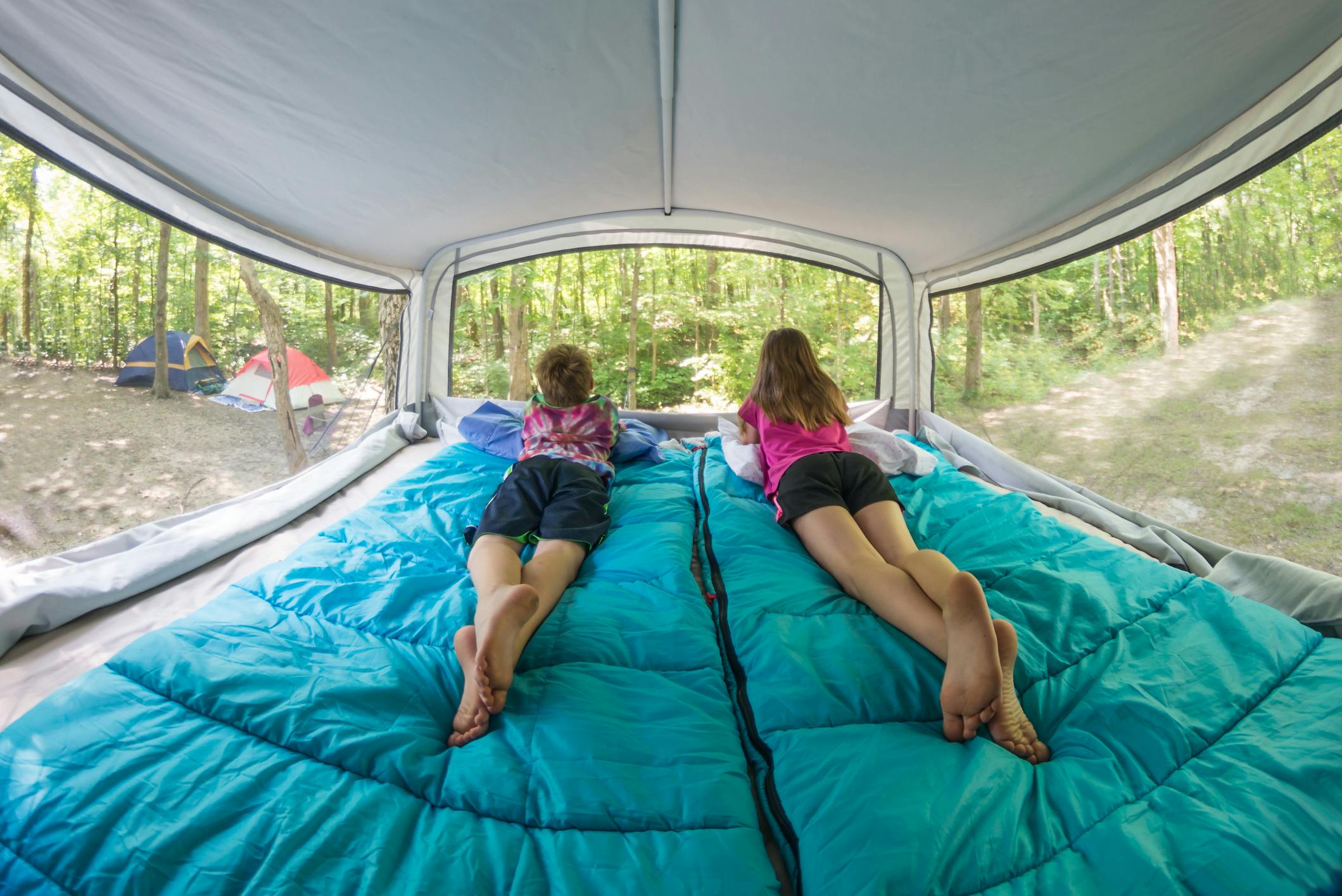 Bare footed children laying on blue sleeping bags in pop-up camper bunk end bed looking out window at surrounding forest trees and campground.