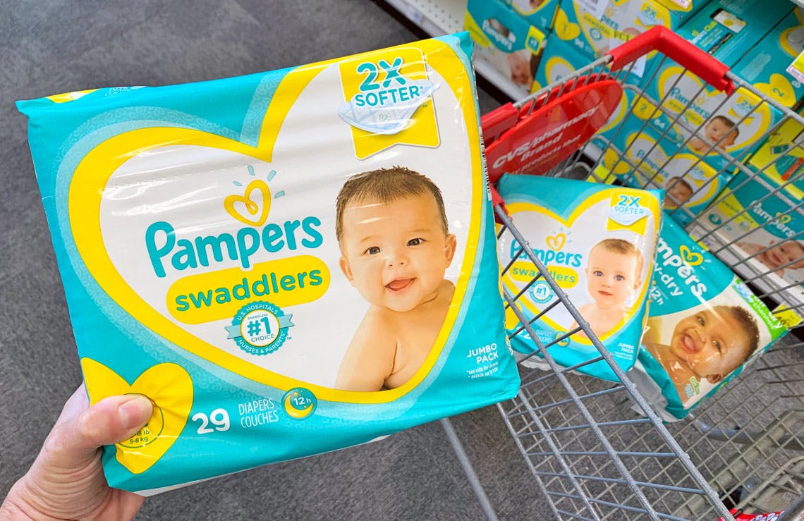 A person's hand holding up a Jumbo Pack of Pamper's swaddlers diapers in front of a shopping cart, with two more packages of diapers, parked in an aisle at CVS.