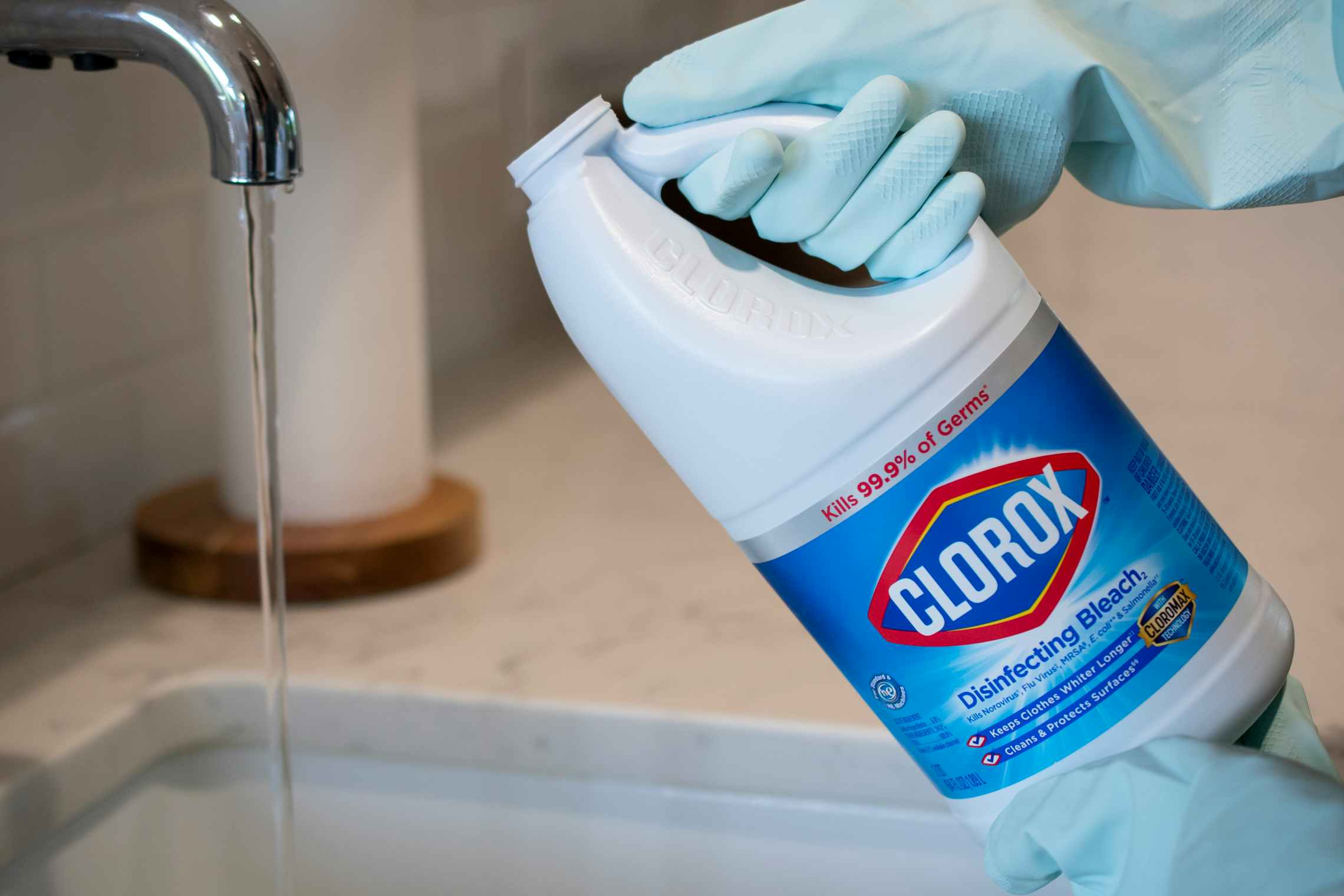 A person holding a bottle of Clorox bleach next to a sink with the tap running while wearing rubber dish gloves.