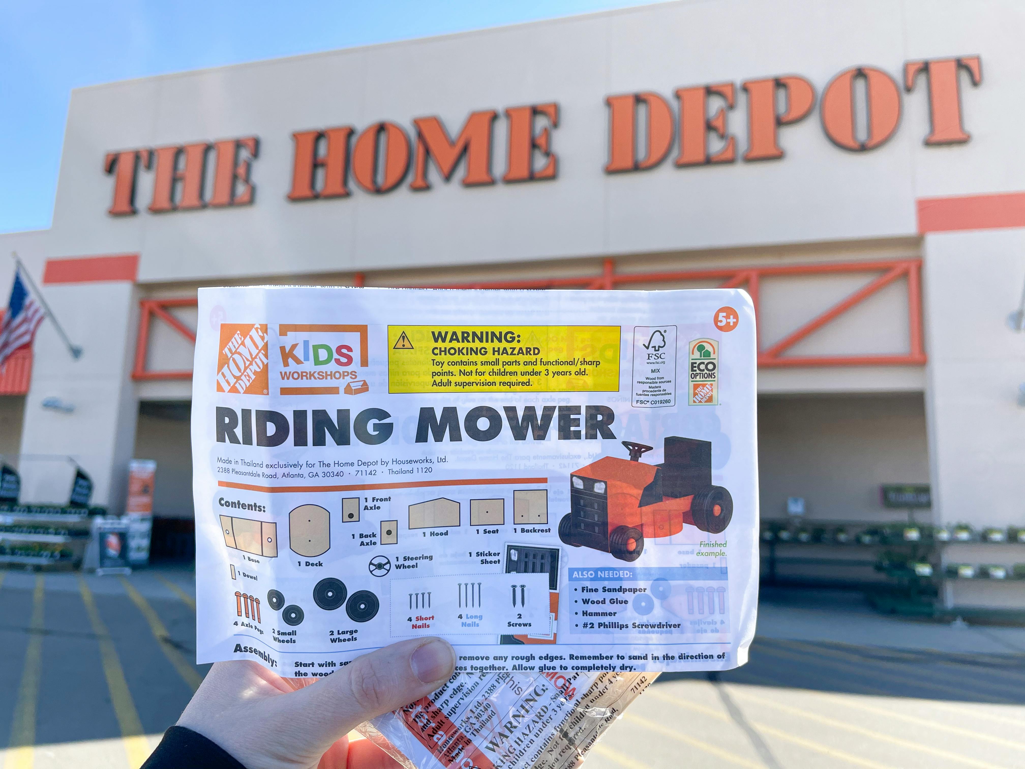 A Home Depot Kids Workshops Riding Mower kit held in front of a Home Depot storefront.