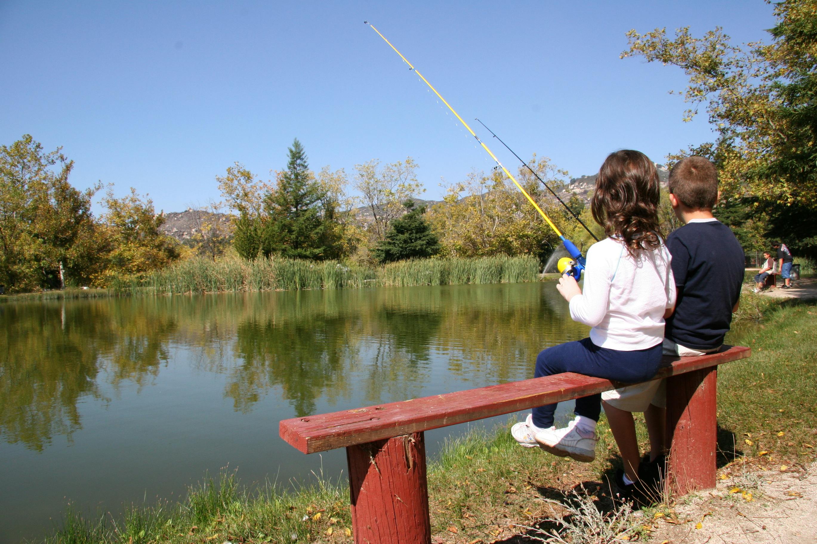 Two kids sitting on a bench holding fishing poles by a lake