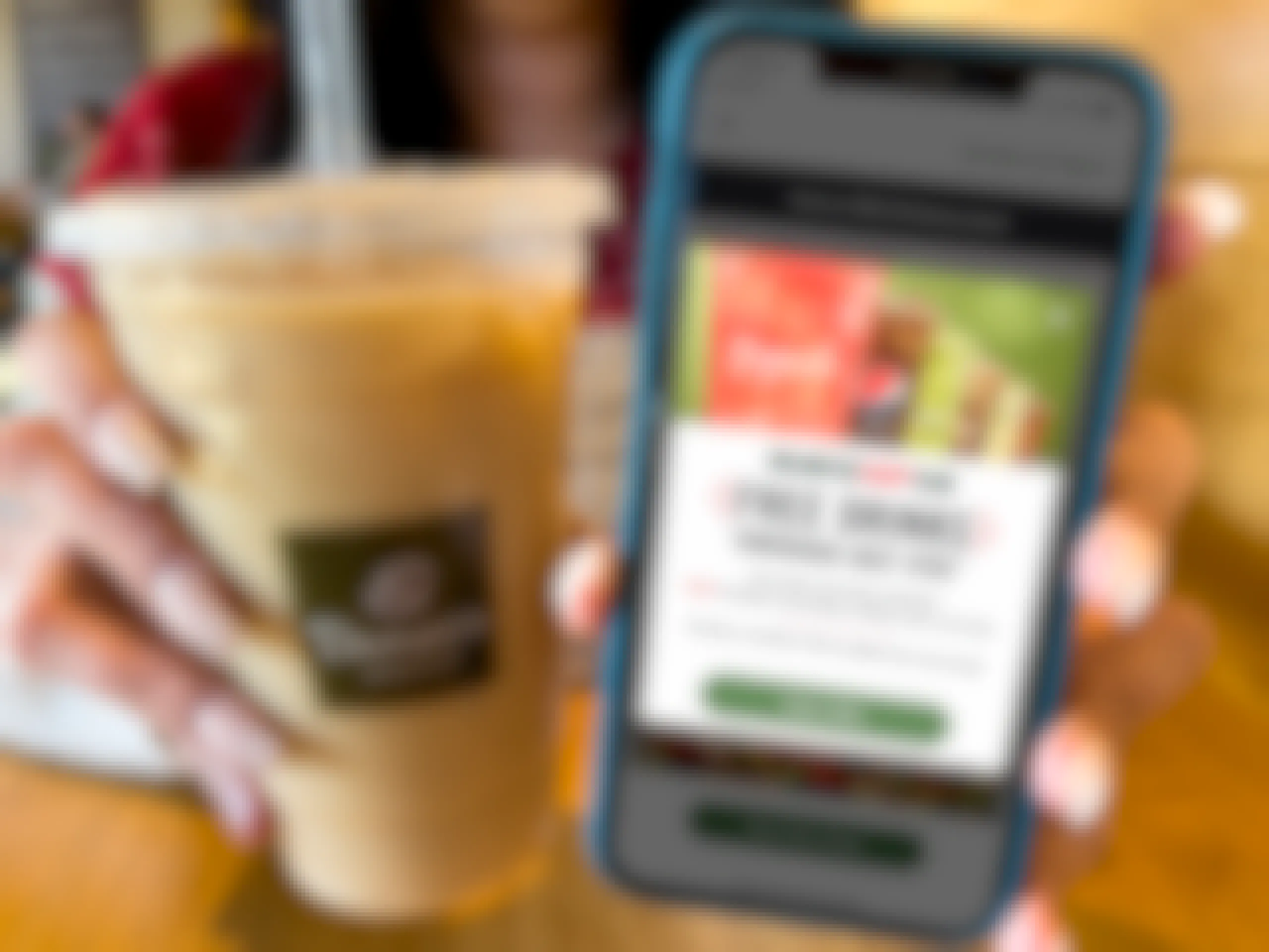 A woman holding an iced coffee in a panera cup next to a cell phone displaying a deal for the unlimited sip club on the panera app.