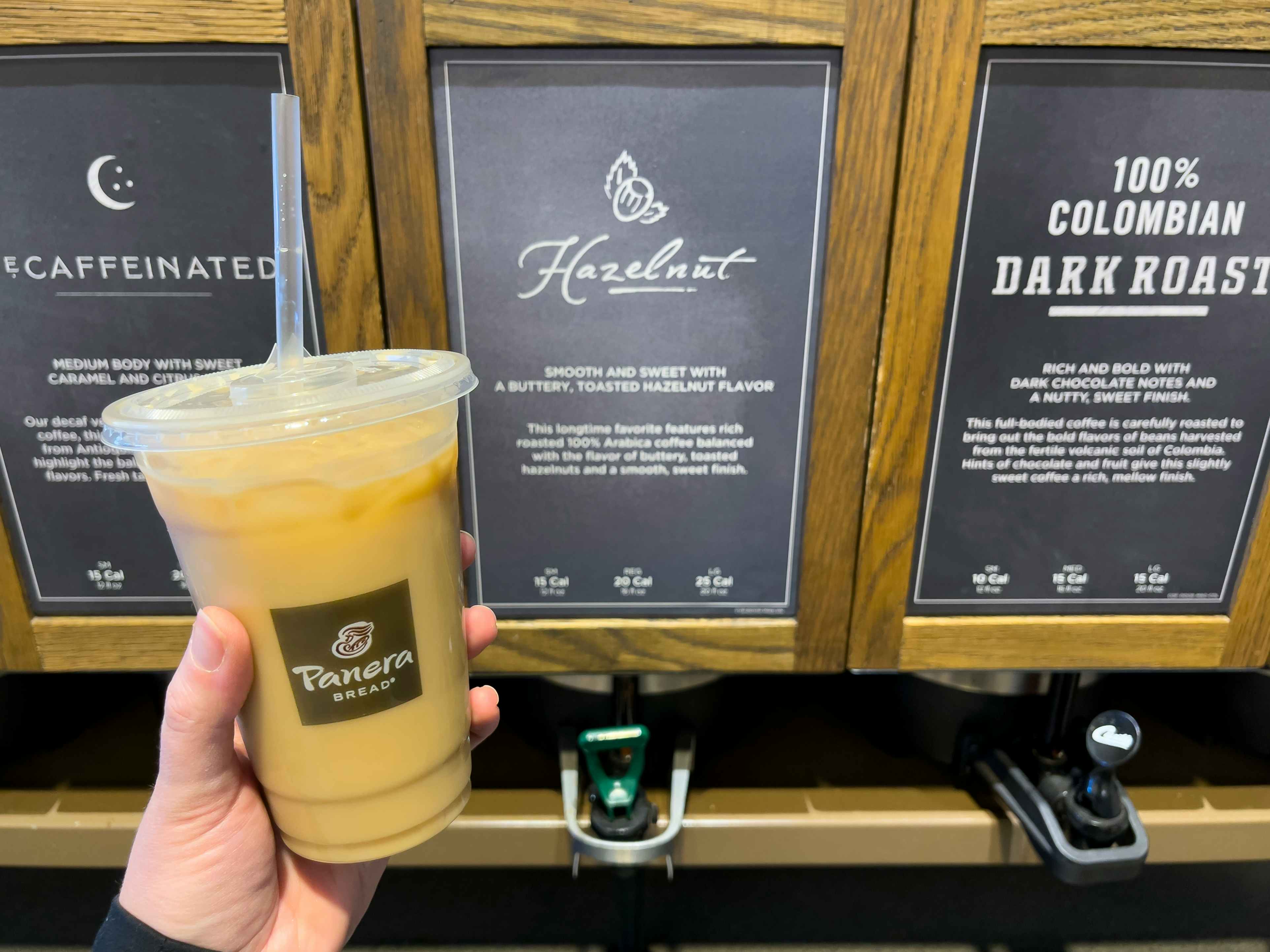 A person holding an iced coffee next to the Hazelnut coffee at Panera Bread.