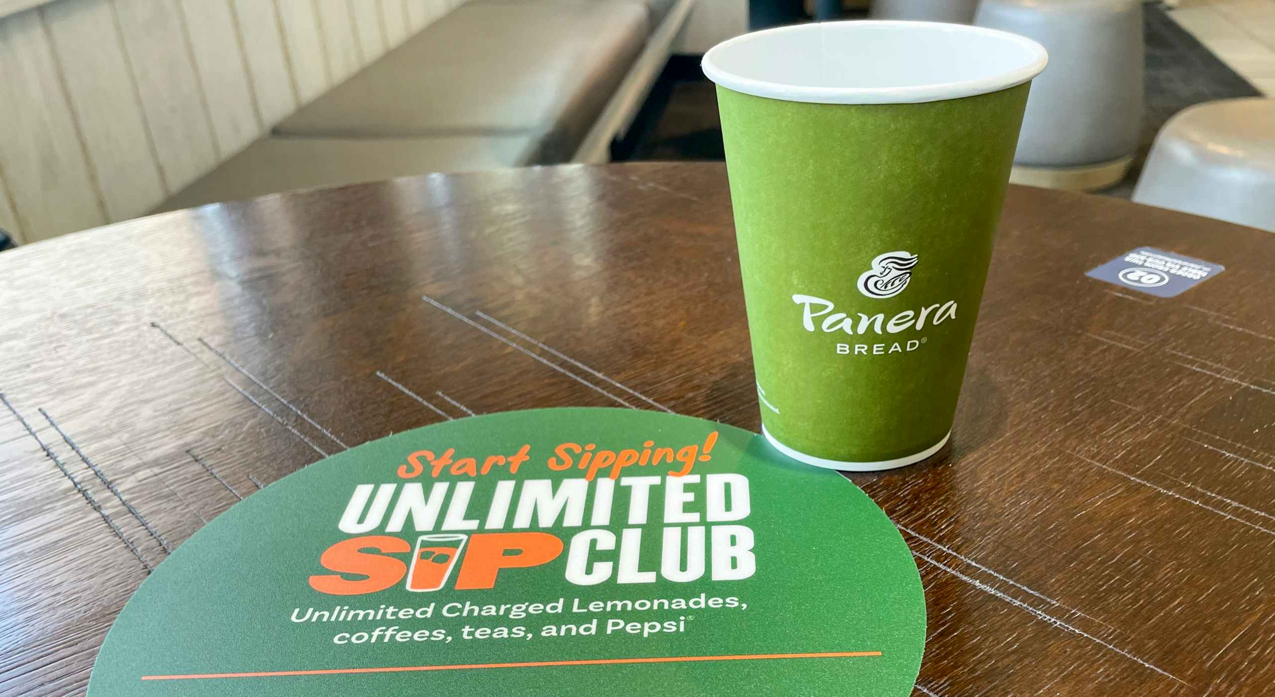 A panera bread cup next to an unlimited sip club sticker on a table.
