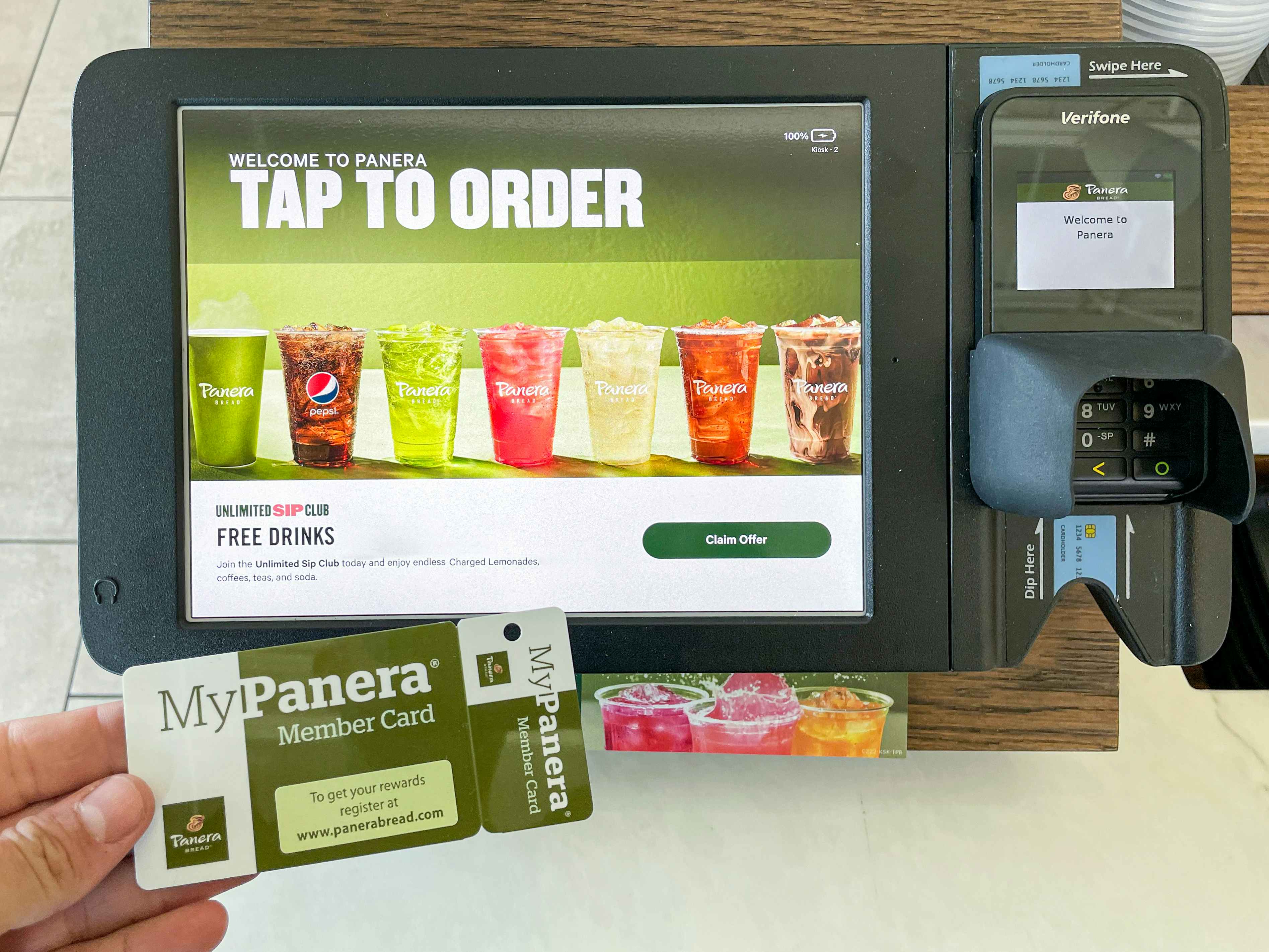 A person's hand holding a MyPanera Member Card next to the POS machine at a Panera counter.