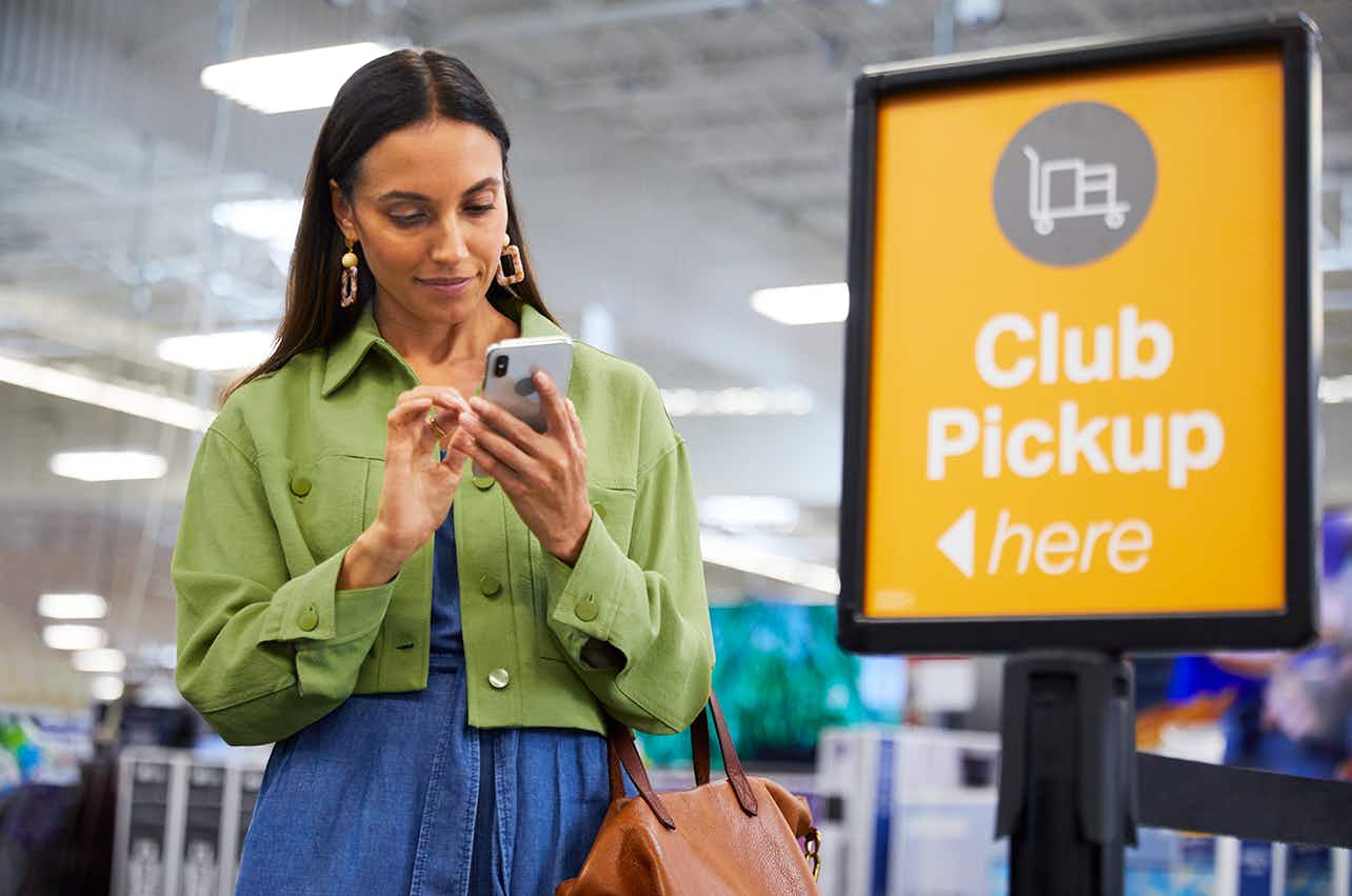 A customer looking at her phone while standing next to a club pick up sign inside Sam's Club