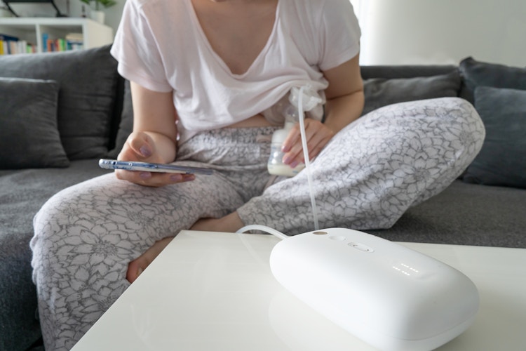 A woman sitting on a couch, using a breast pump and looking at her phone.