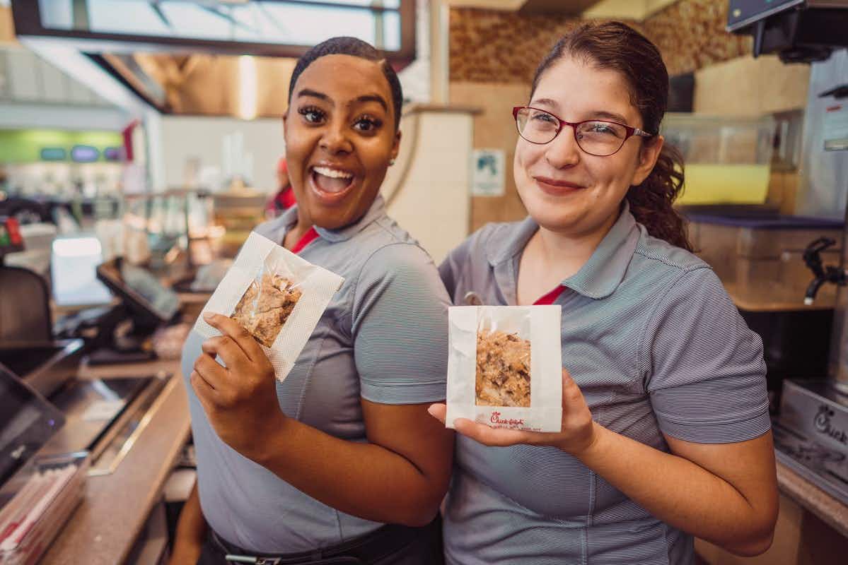 Two Chick-fil-A employees standing behind the counter inside a Chick-fil-A, holding chocolate chunk cookies.