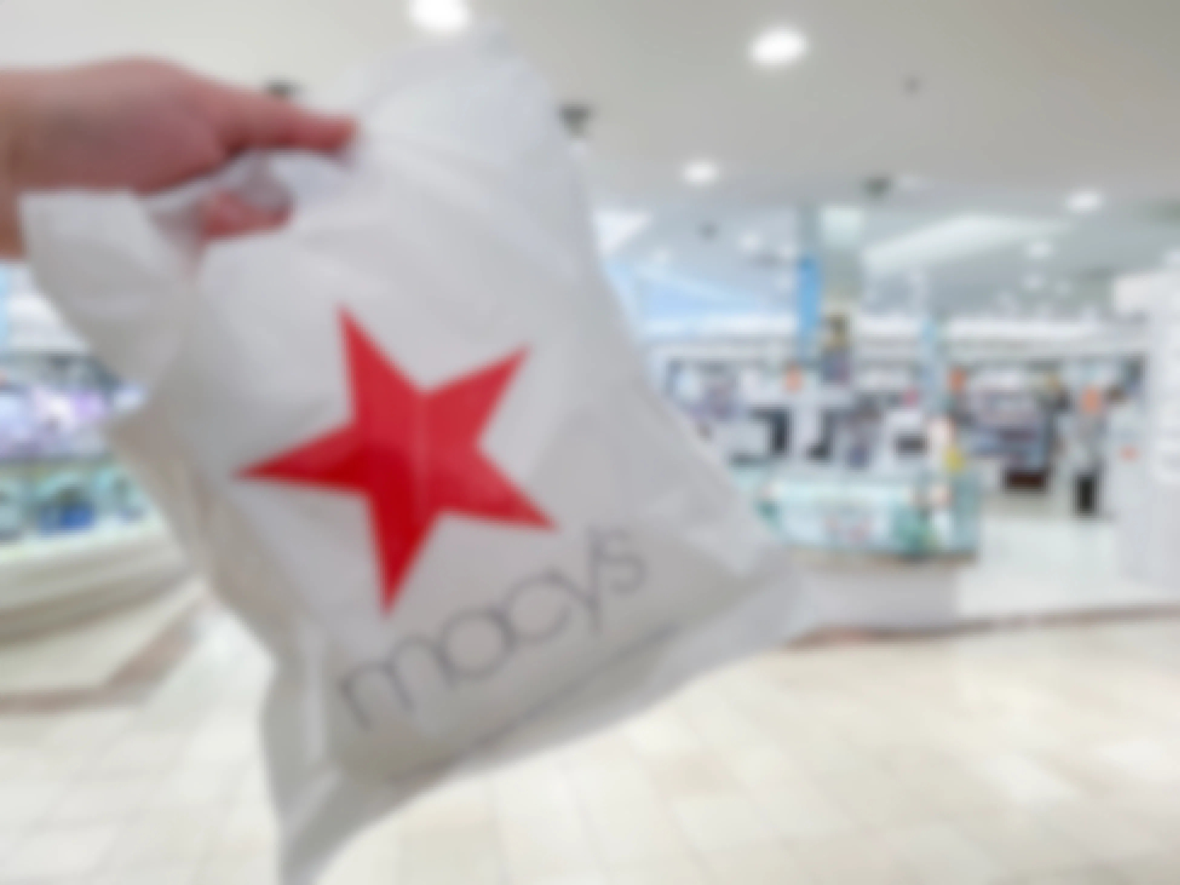 A Macy's shopping bag held in front of the cosmetic section inside Macy's.