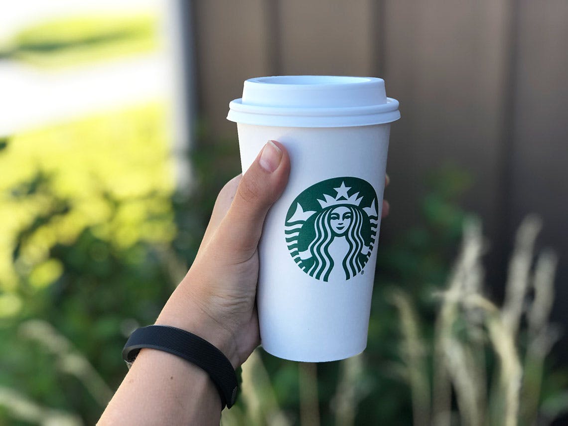 A person's hand holding up a hot Starbucks coffee while outside.