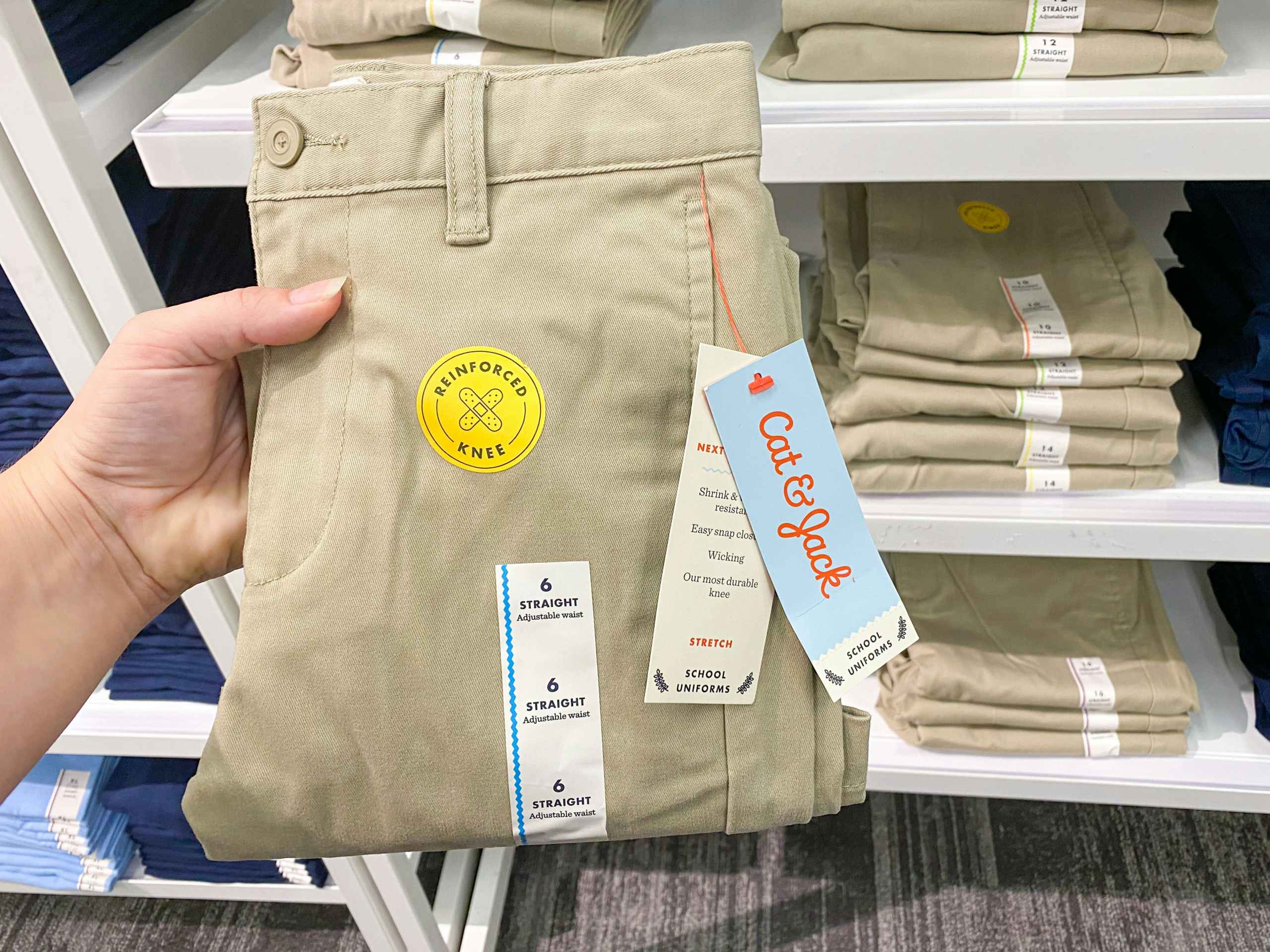A person's hand holding up a pair of Cat & Jack kids school uniform slacks in front of a shelf of more of the same product at Target.