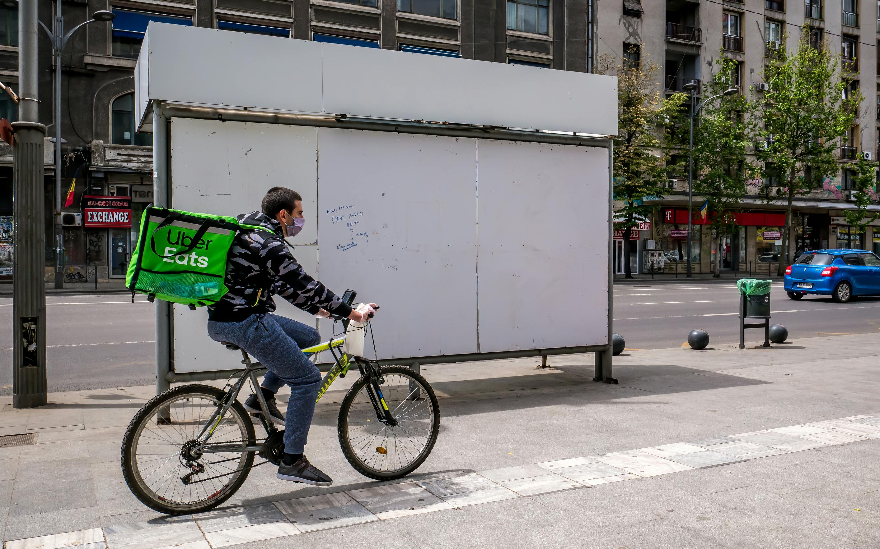 An Uber Eats employee, wearing an Uber Eats cooler delivery backpack, riding a bicycle down a sidewalk.