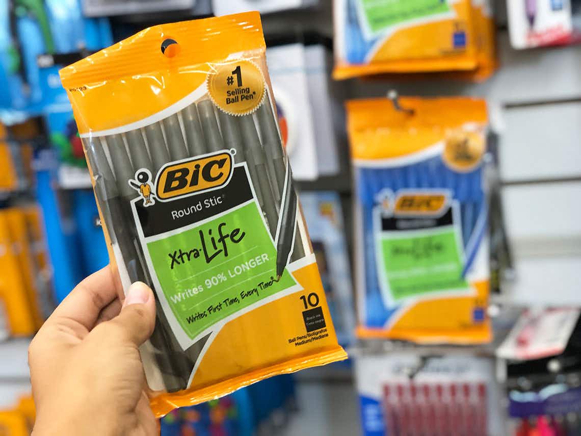 A person's hand holding up a package of BIC pens in front of a wall displaying more pens inside Dollar Tree.