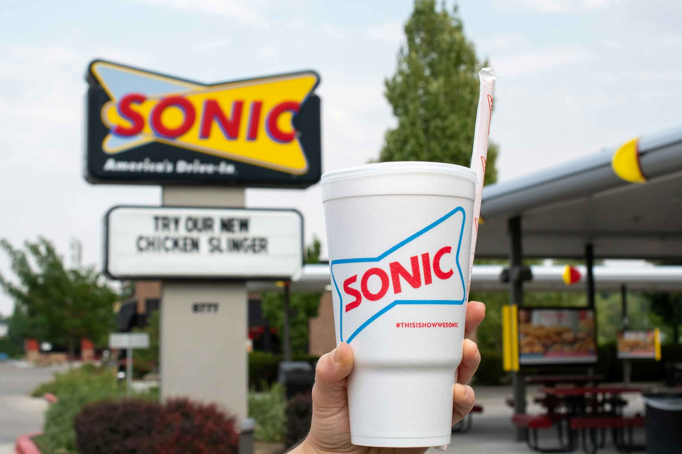 A person's hand holding up a Sonic drink cup and straw in front of a Sonic sign next to the drive-up parking spots.