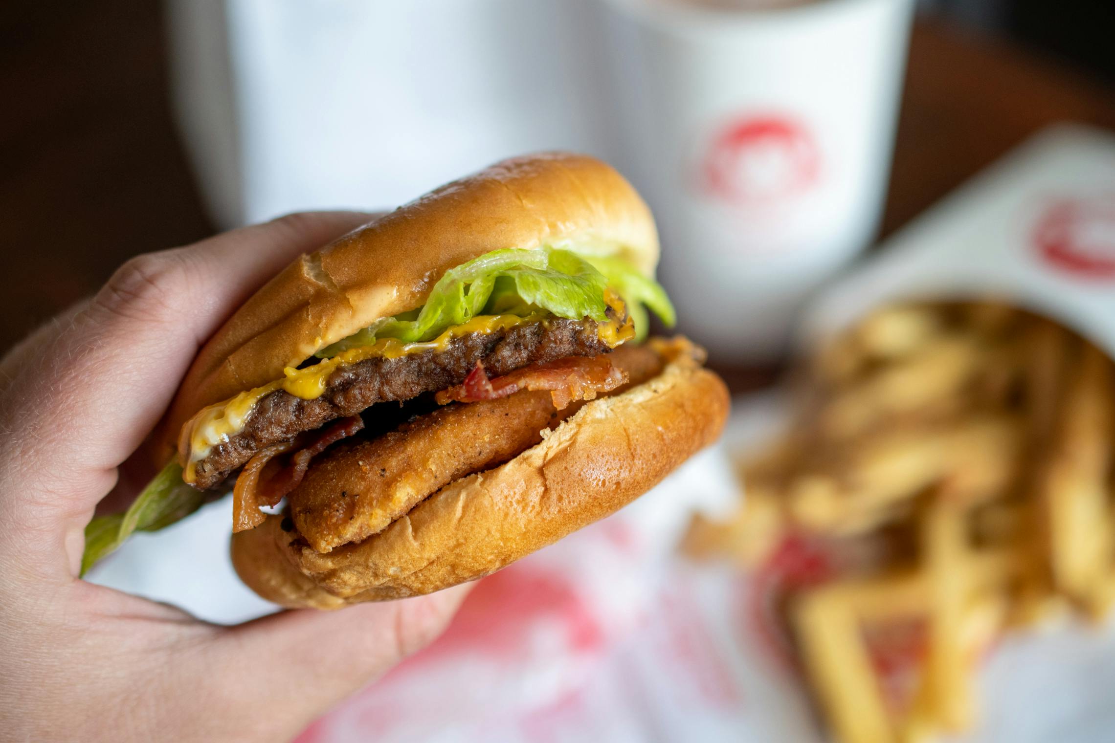 A person holding a Wendy's burger with fries and a frosty out of focus on the table in the background.