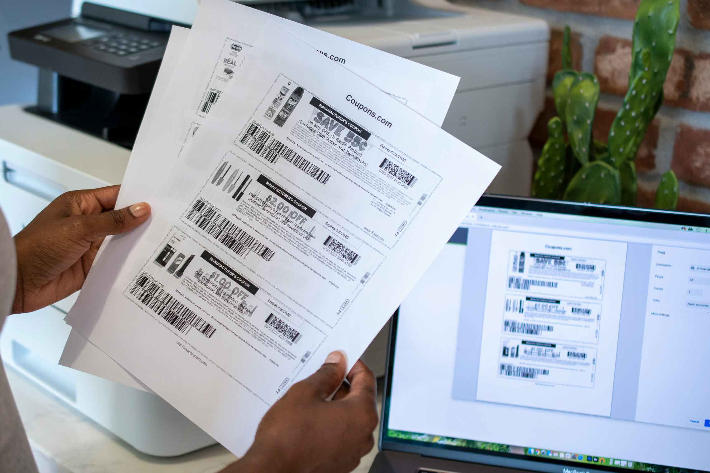 A person holding black and white printed coupons near a printer and computer.