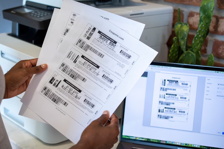 A person holding black and white printed coupons near a printer and computer.