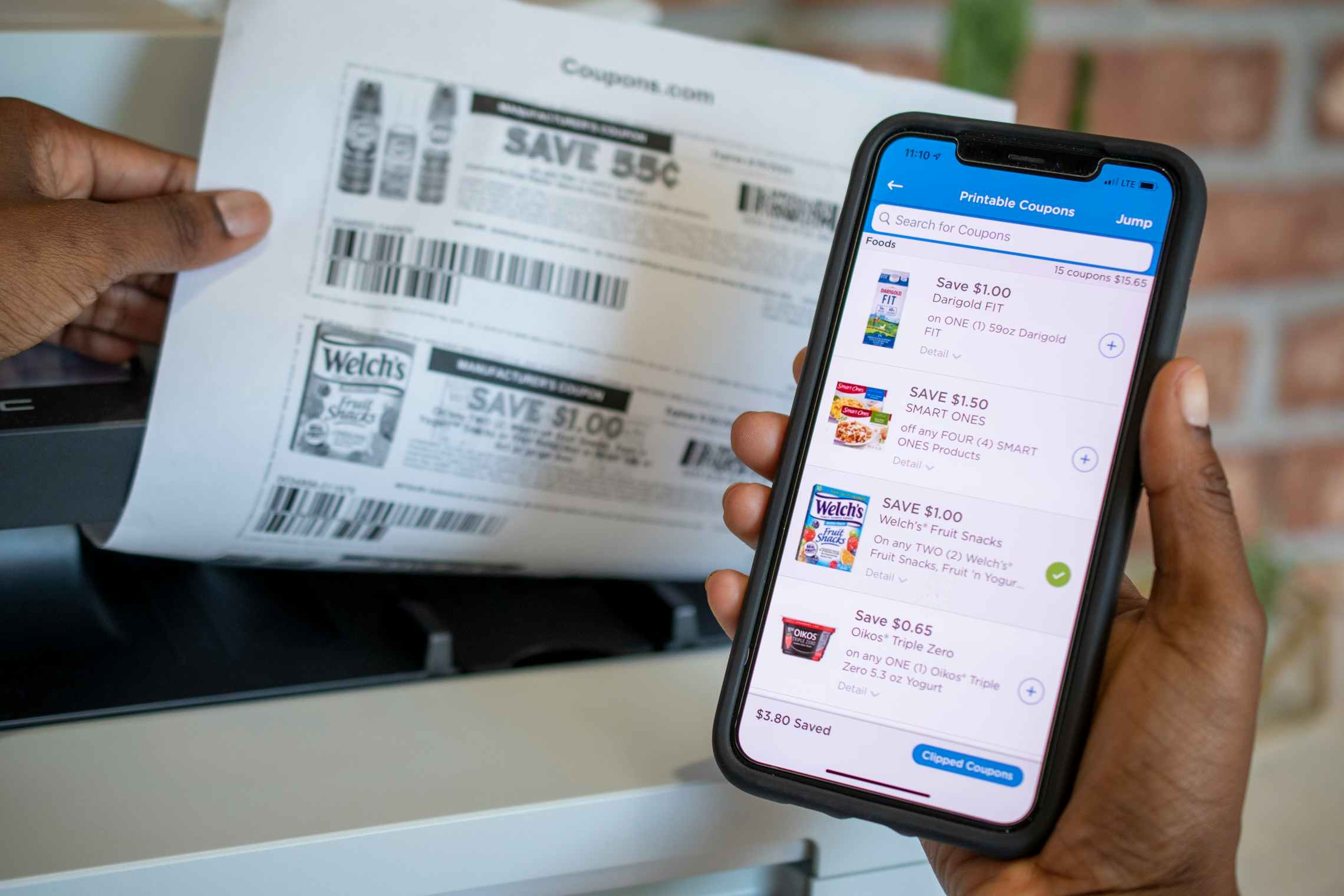 A person's hands, one holding up an iPhone with the Coupons.com mobile app open to the Printable Coupons page, and the other hand taking a sheet of paper with coupons printed on it out of a printer.