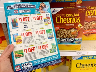 A person holding a box of General Mills cereal with six coupons on the back.