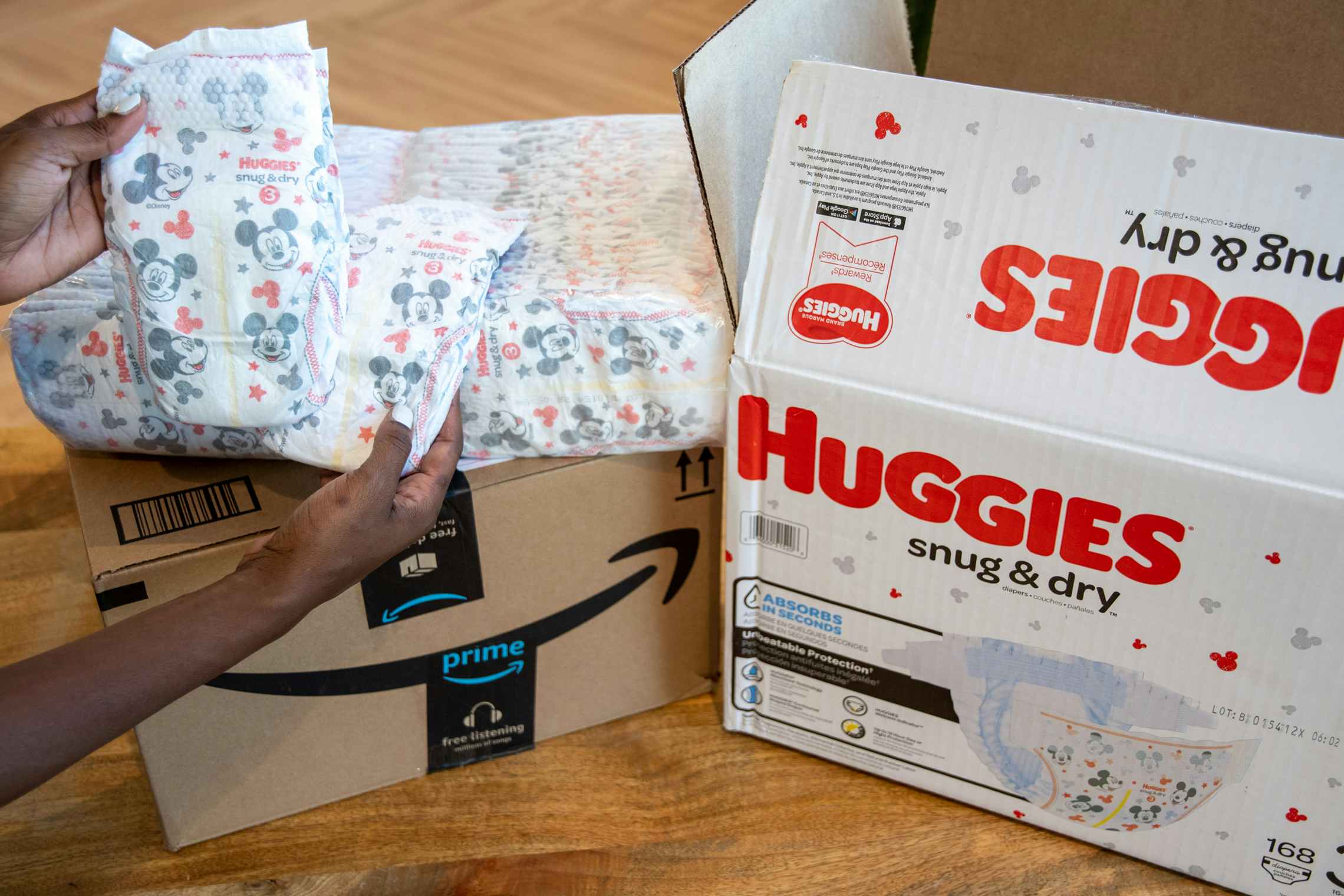 A person's hands holding up a Huggies diaper next to the diaper box and an Amazon delivery box.