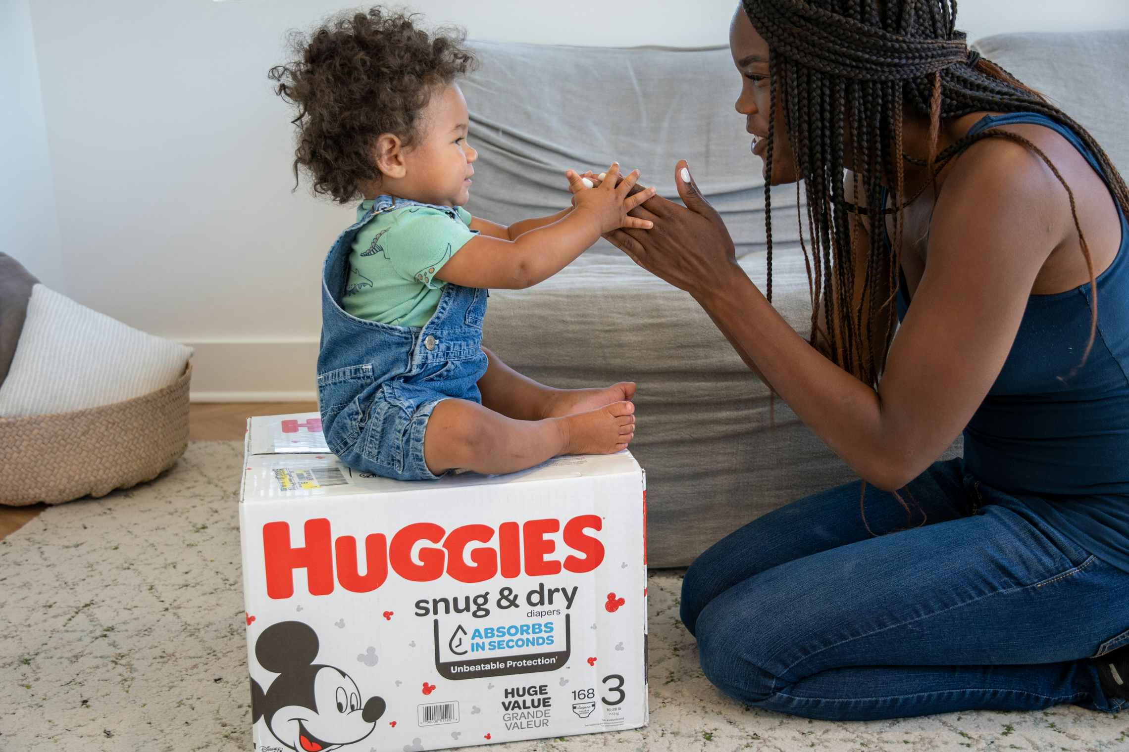 A baby sitting on a Huggies diaper box playing with his mommy.