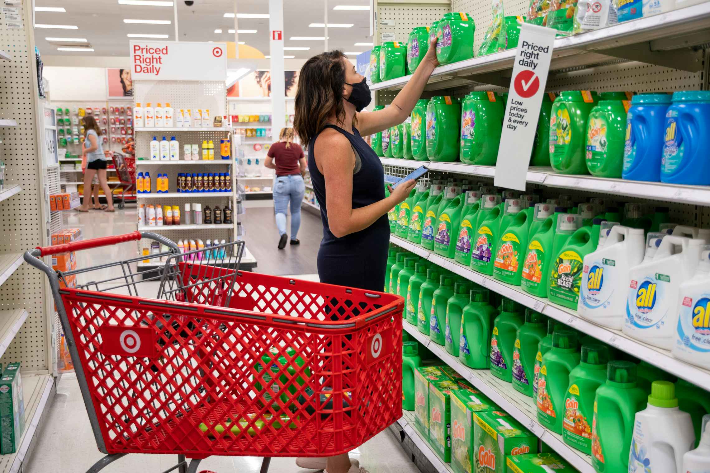A woman standing in the laundry detergent aisle at Target, reaching to grab a container of Gain off of the top shelf. Next to her is a Target shopping cart with other laundry products sitting in it.