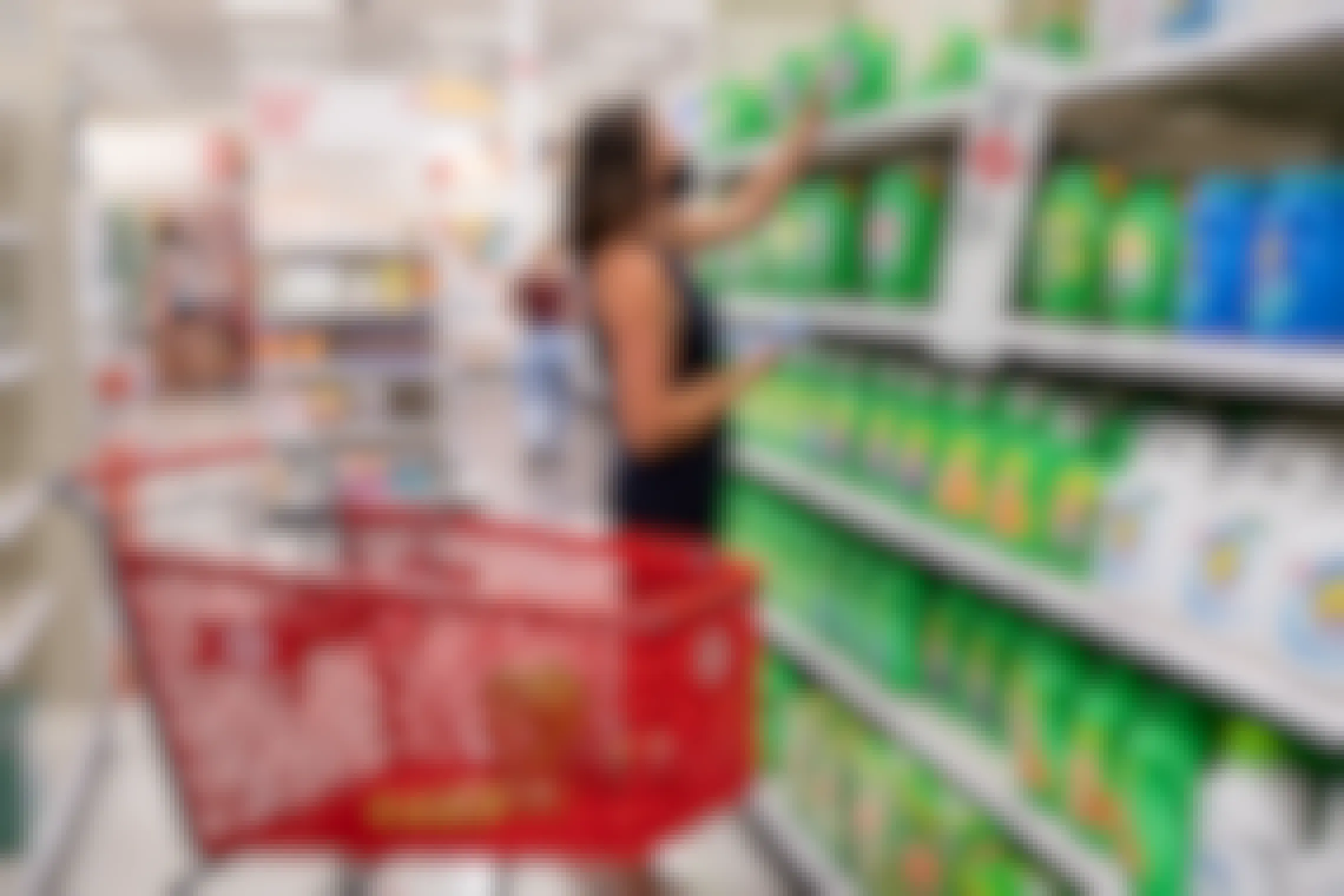 A woman standing in the laundry detergent aisle at Target, reaching to grab a container of Gain off of the top shelf. Next to her is a Target shopping cart with other laundry products sitting in it.